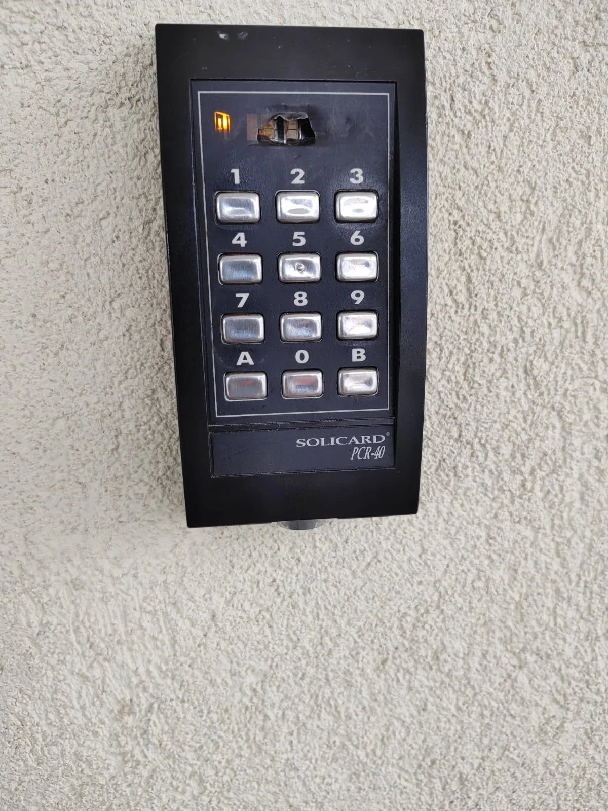 Elektronisk adgangskodepanel festet til en vegg, med knapper merket med tallene 0-9 og bokstavene A og B, og en liten skjerm øverst.
