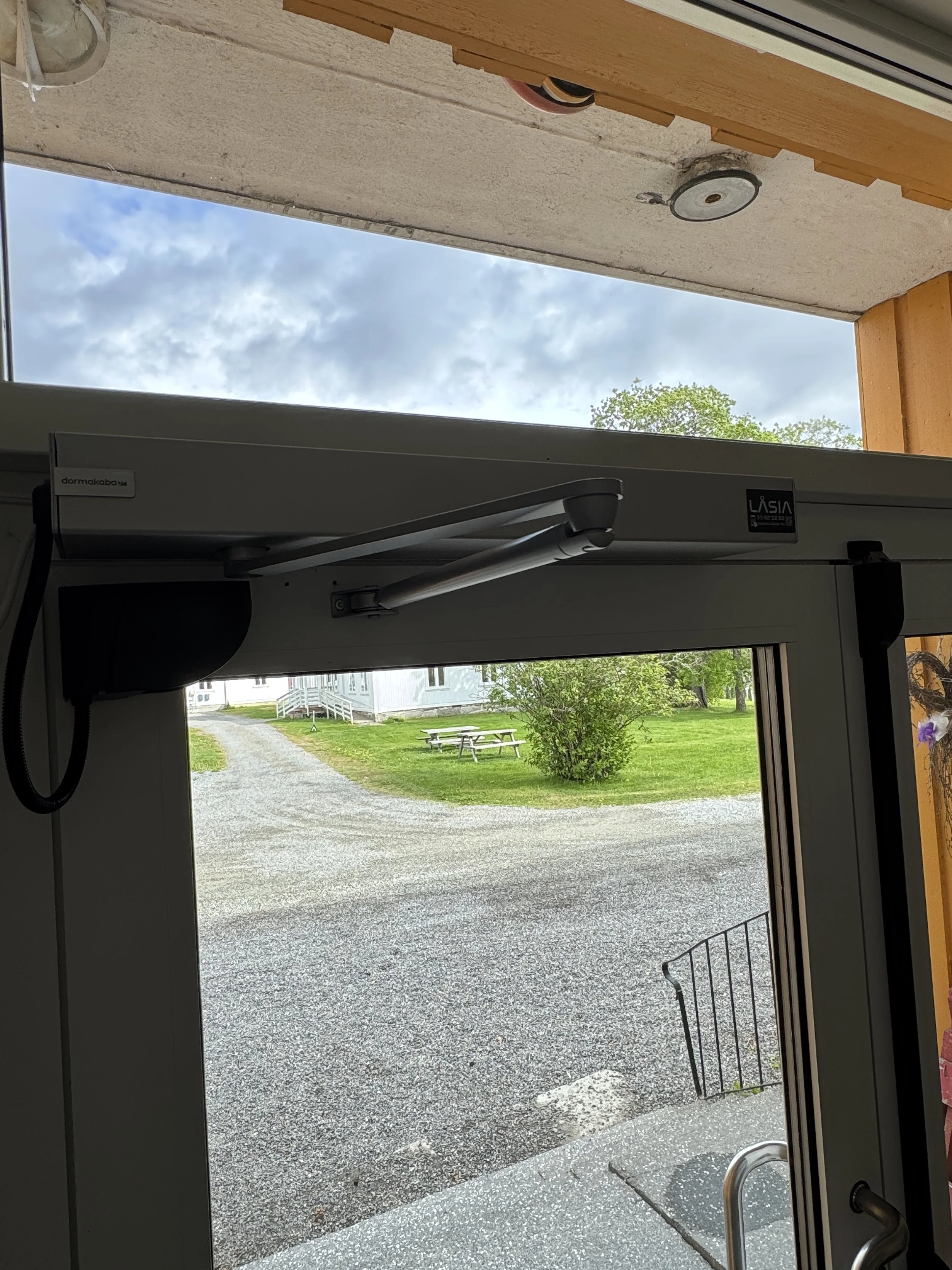 Utsikt fra en inngangsportal mot en grusvei, grønne plener, en busk og noen benker, med en bygning i bakgrunnen.