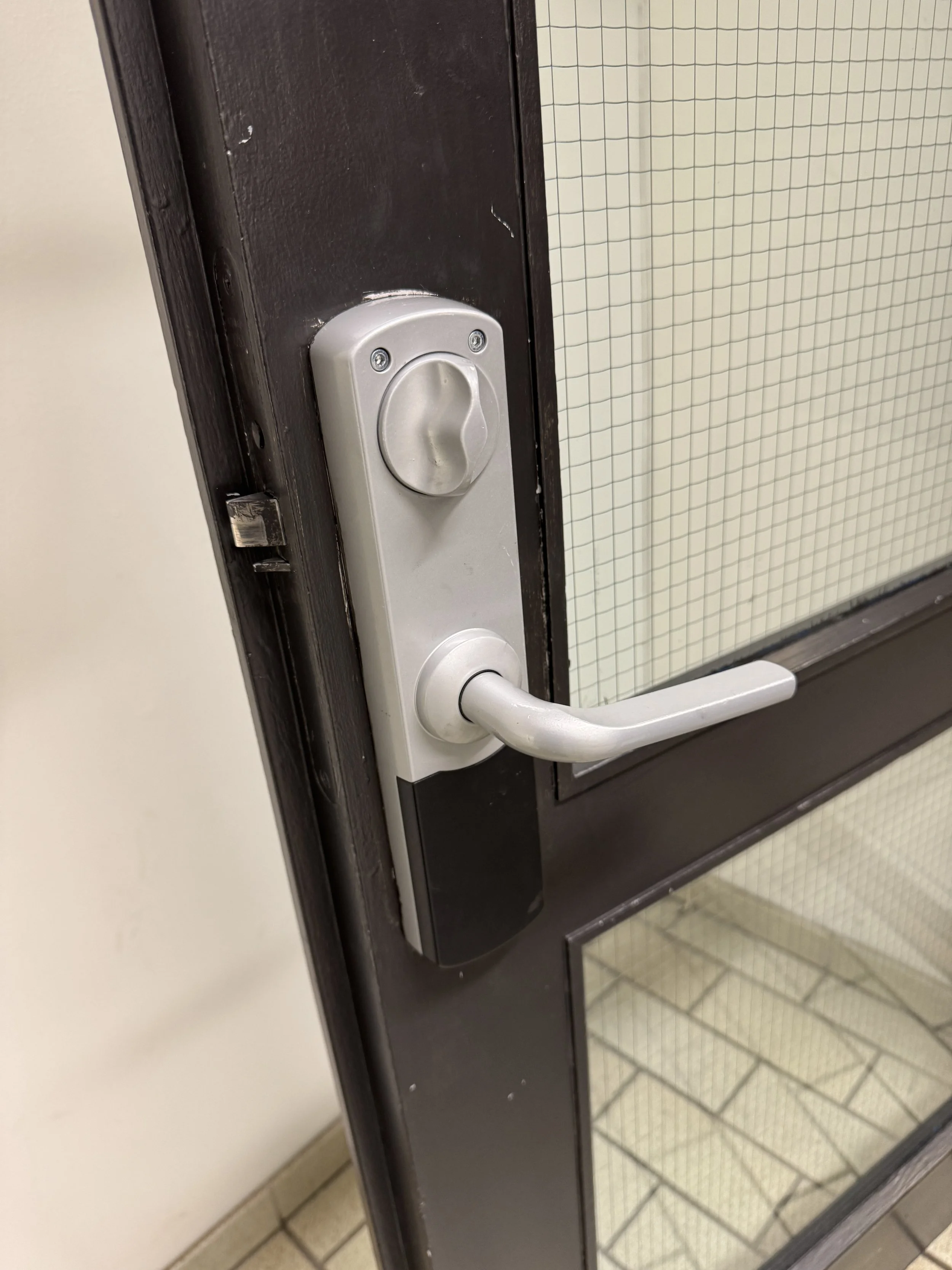 Nærbilde av en dør med en hvit dørhåndtak og lås, lager av svart metall, og et glassfelt med rutete mønster.