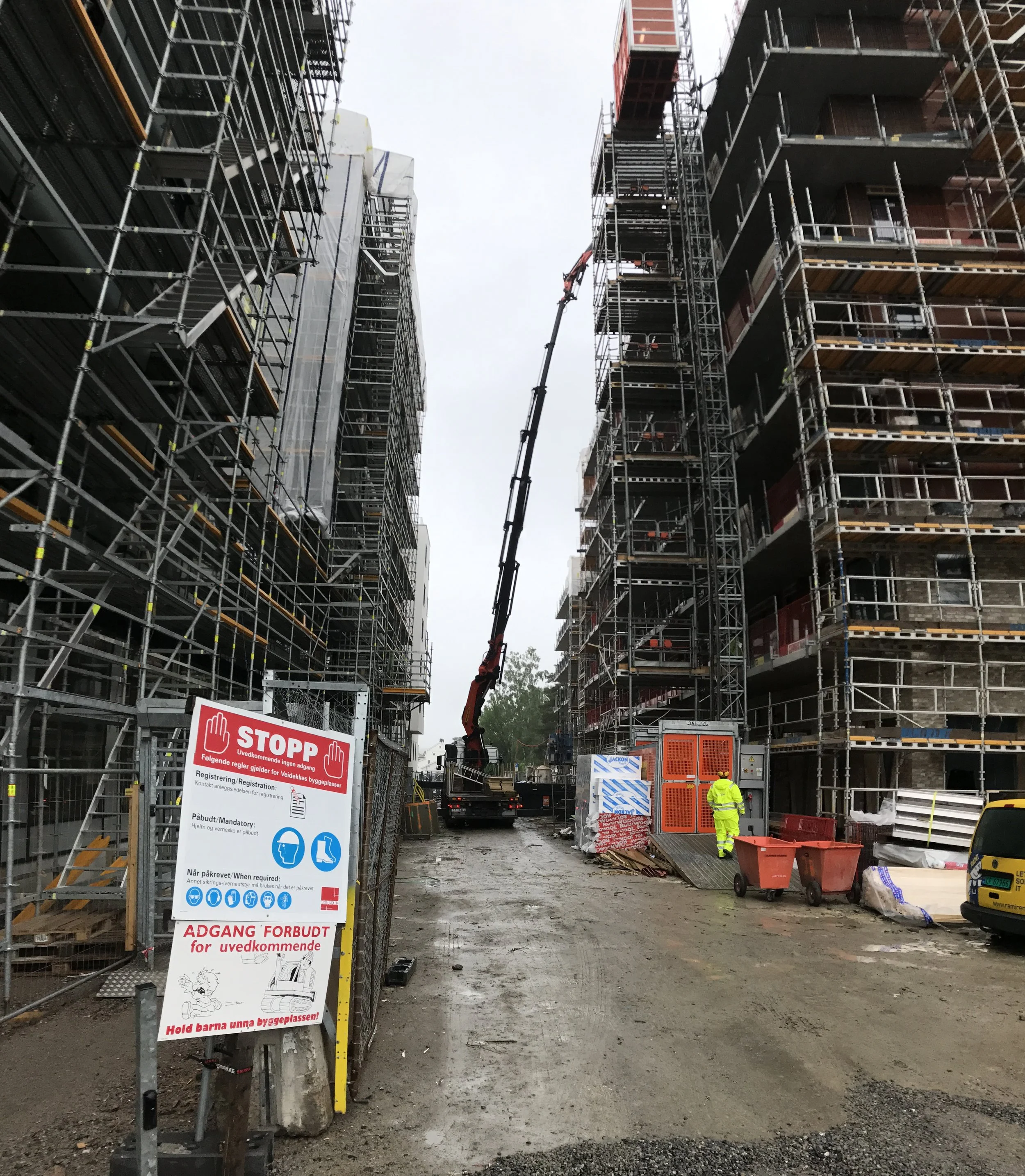 Byggeplass med høye bygninger omgitt av stillas, en person i høy synlighet klær, og byggemateriell, samt skilt som varsler om trygghet, i Norge.