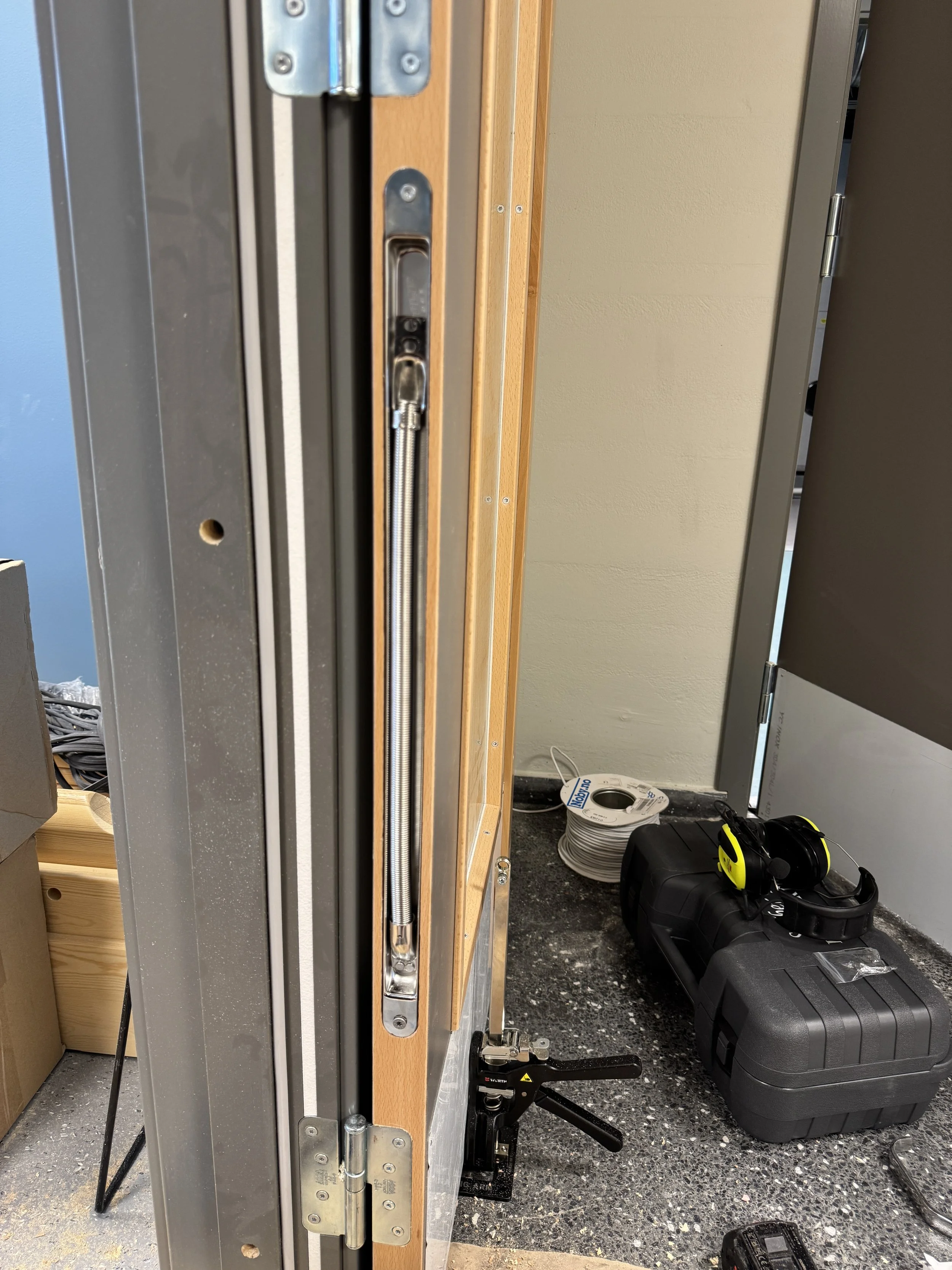 Inne i et byggefellesrom med en dørkarm i metall, en dreibar låsearm, en trapp på gulvet, en oppspenning med hjul og verktøy, en sort hylse med hørselvern på, og en trådspole på gulvet.