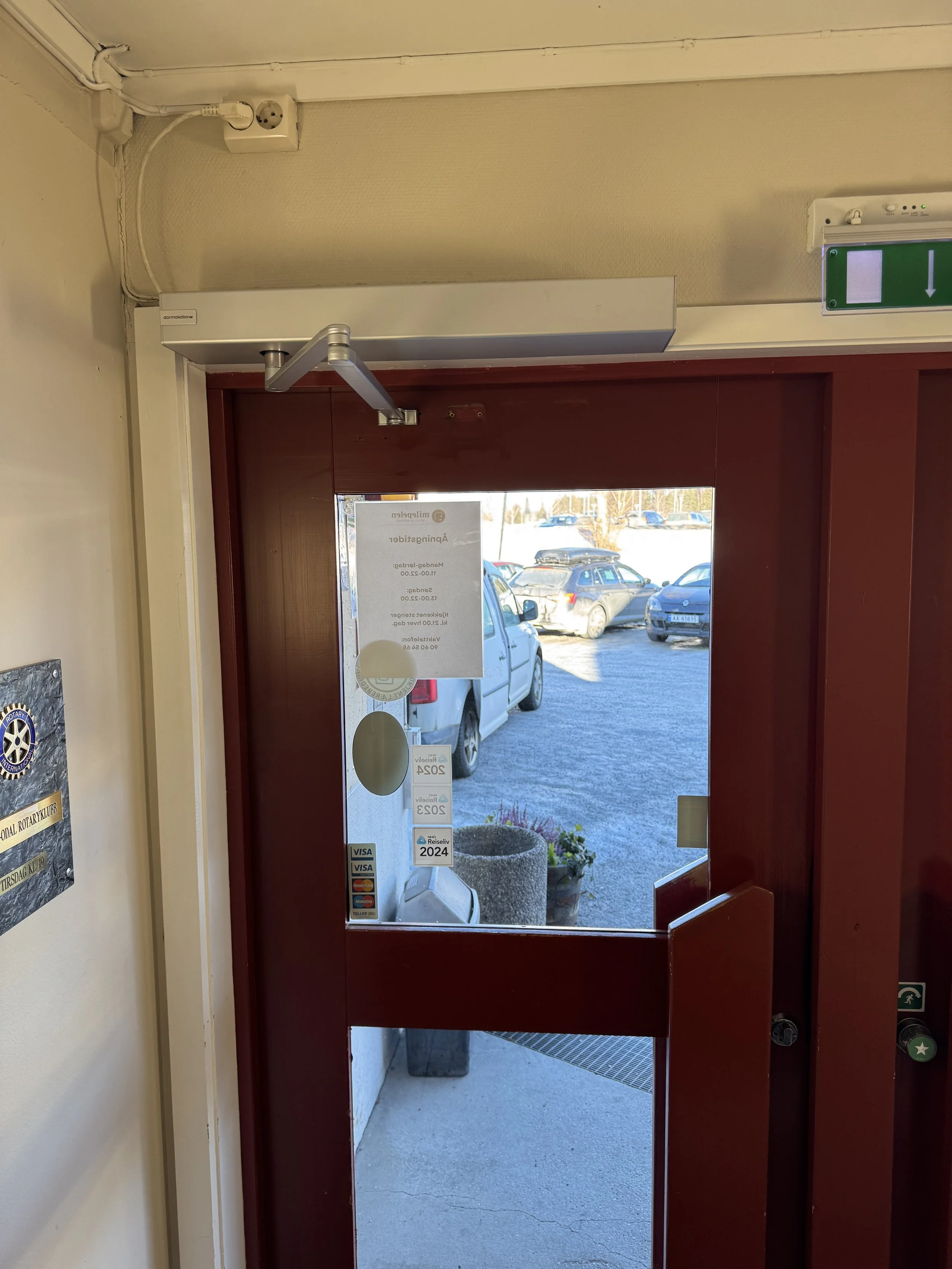 Utsikt gjennom en dør mot en parkeringsplass med biler, og en stor blomsterpotte utenfor. En oppslagstavle er festet på døren, og det er et utgangsskilt over døren.