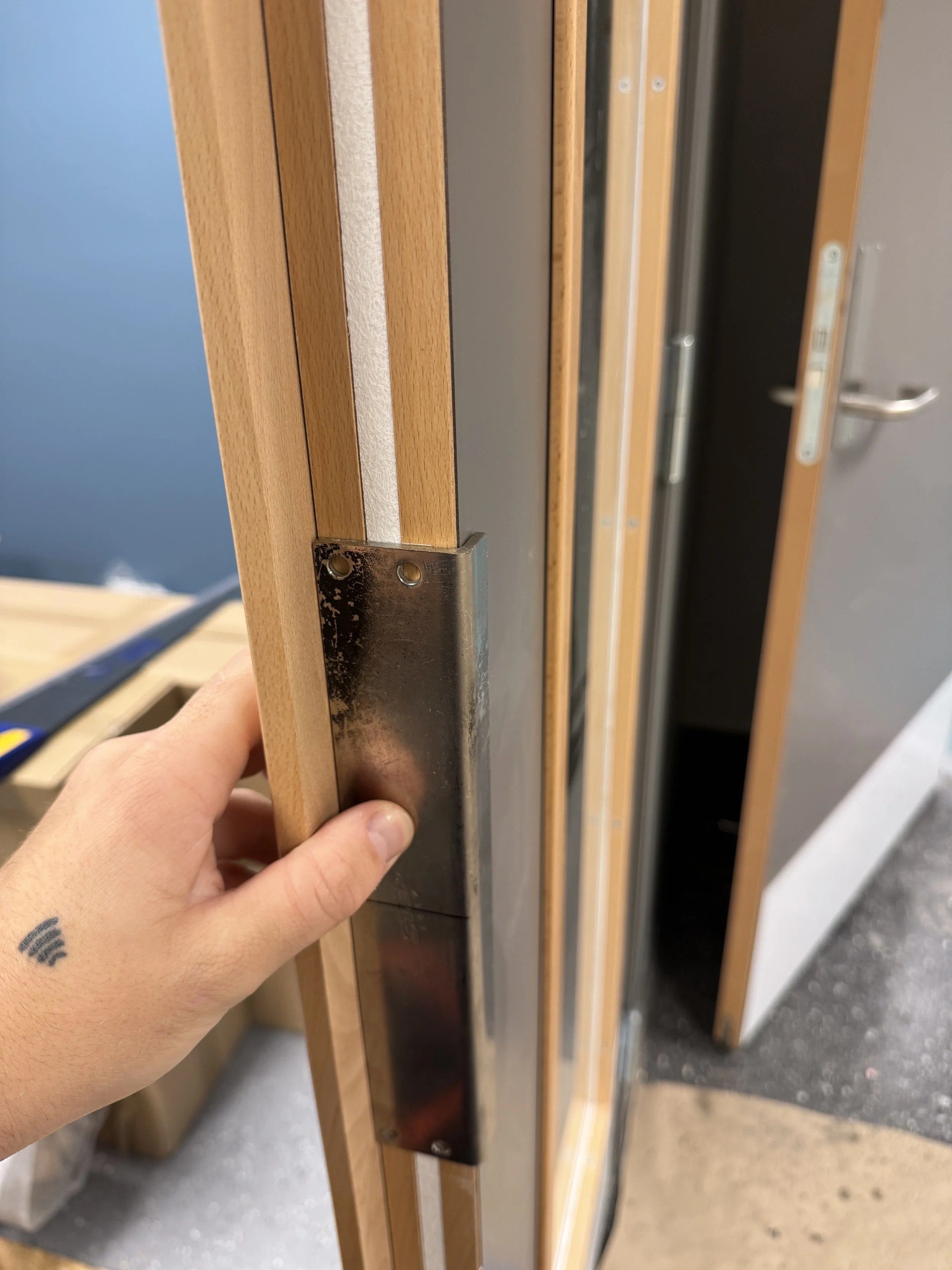 Nærbilde av en hånd som holder en metallbeskytter foran en treramme. Det er flere treverk og metallrammer i bildet.