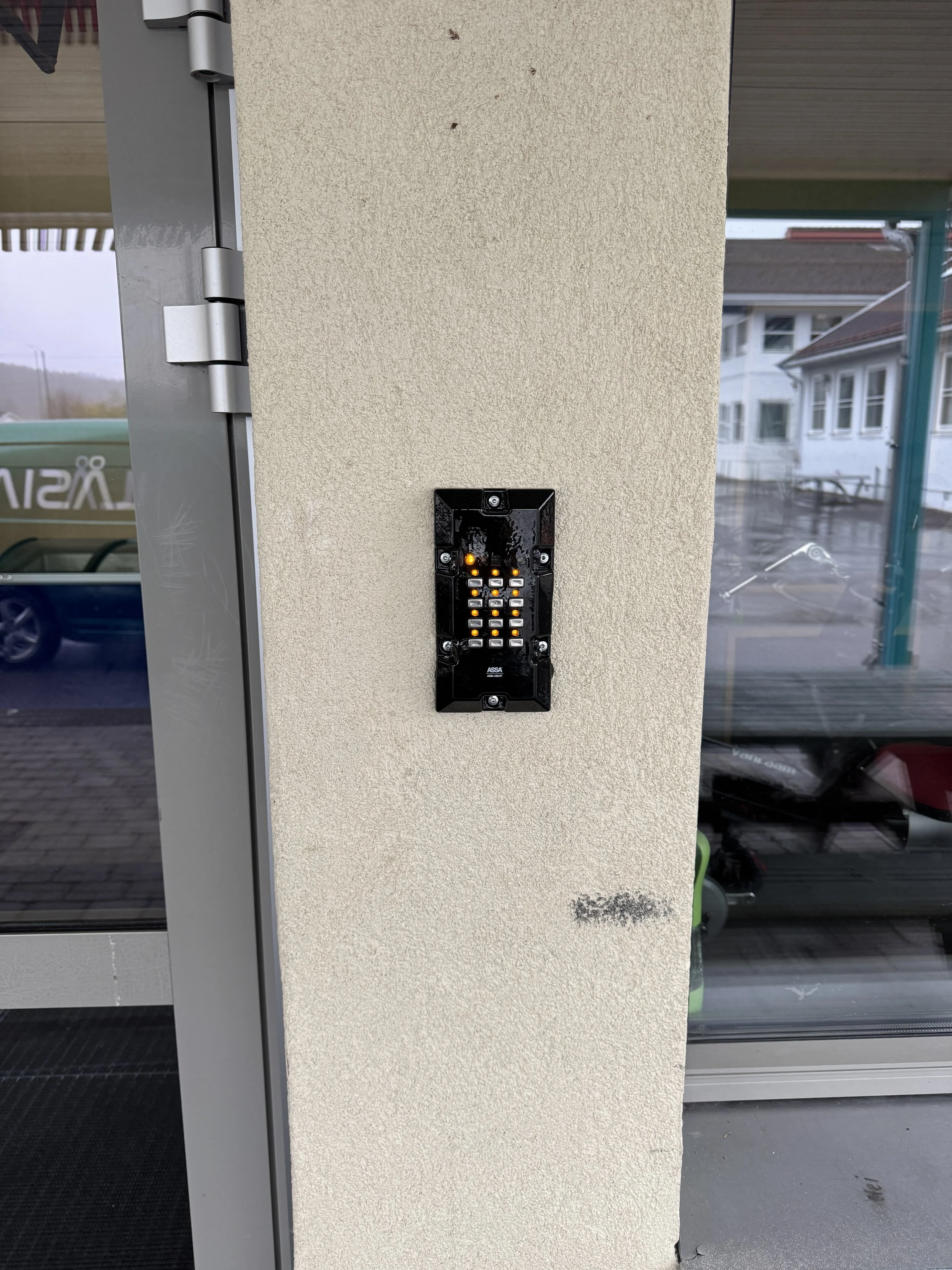 Våpenfrigjøringsanlegg på en vegg ved inngangsdør, med flere små knapper og lys. Utenfor er det regnvær og et bygget utendørs.