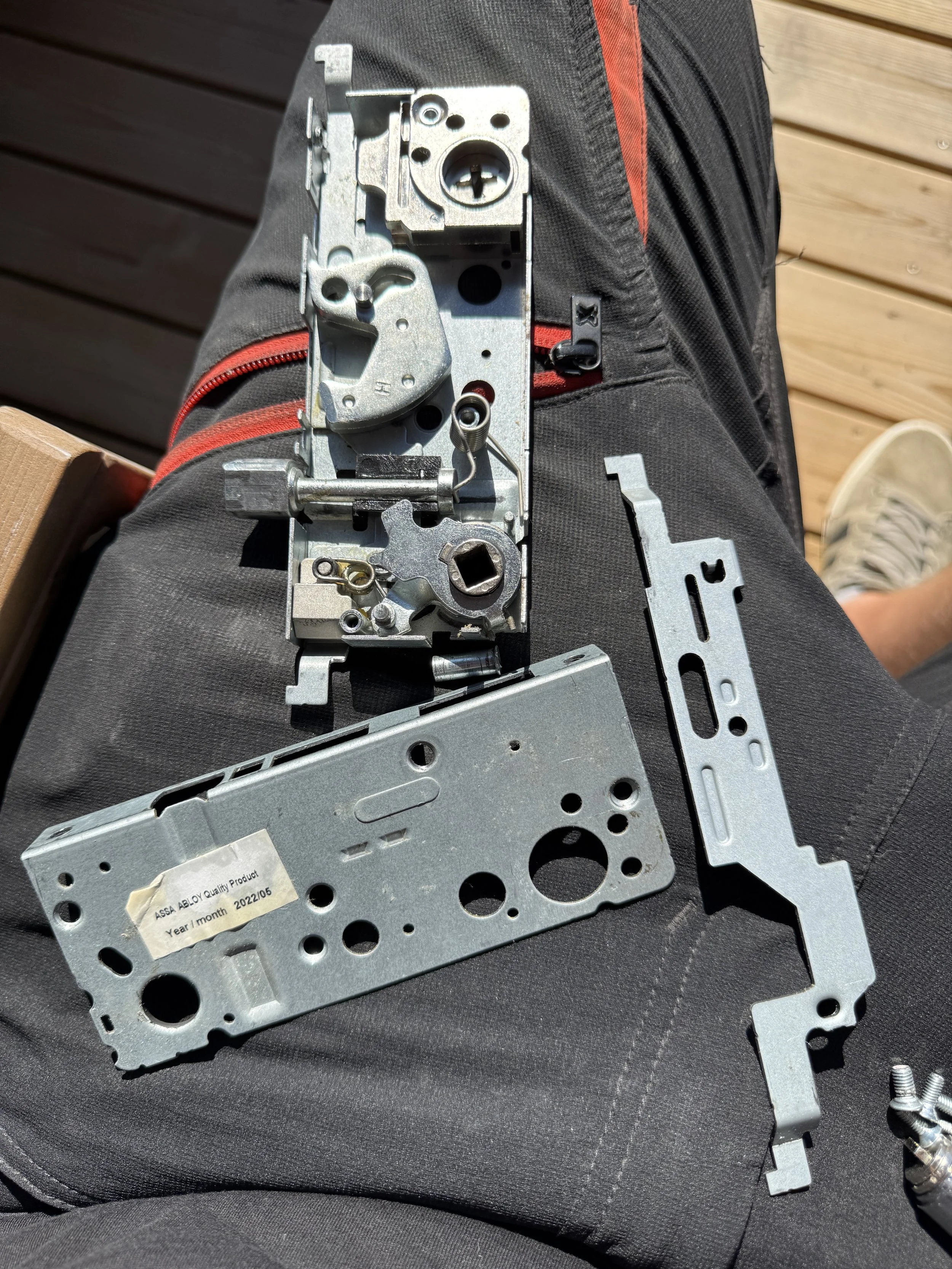 Utsikt av metallkomponenter og deler som ligger på en persons lår, med en utendørsbakgrunn av treplanker og en sko i bakgrunnen.