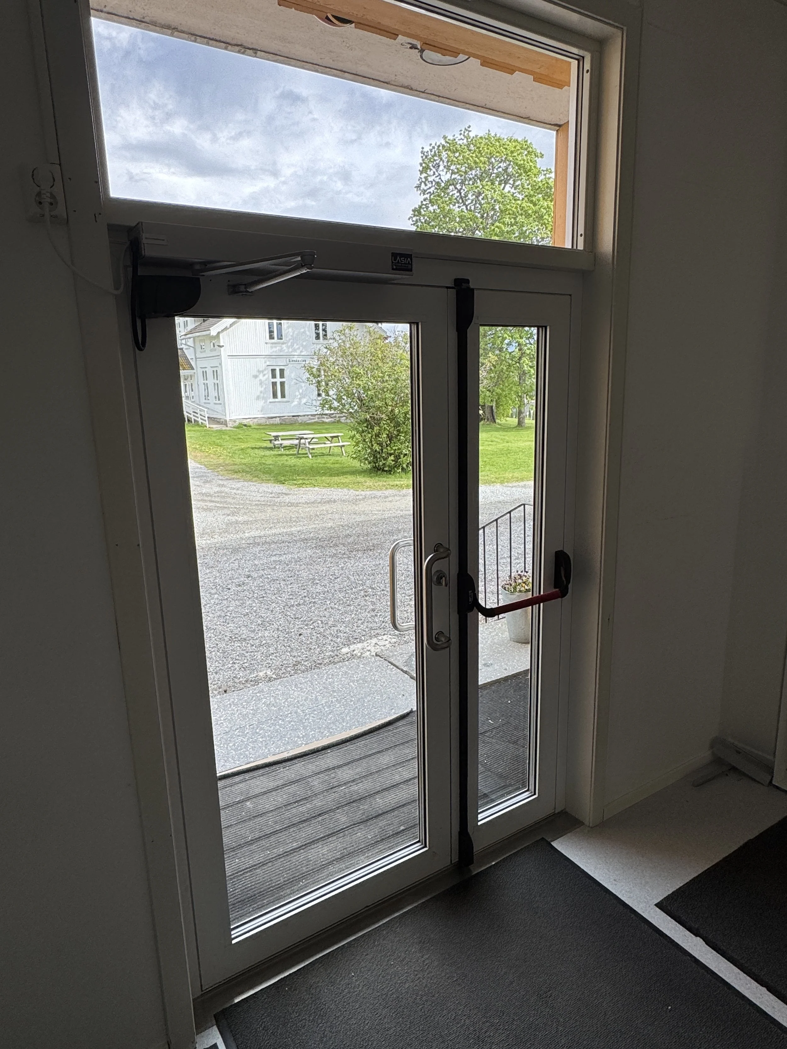 Nærbilde av en utgangsdør med glass og rullestolvennlig døråpner, som viser utendørs gårdsplass med gress, trær og en hvit bygning i bakgrunnen.