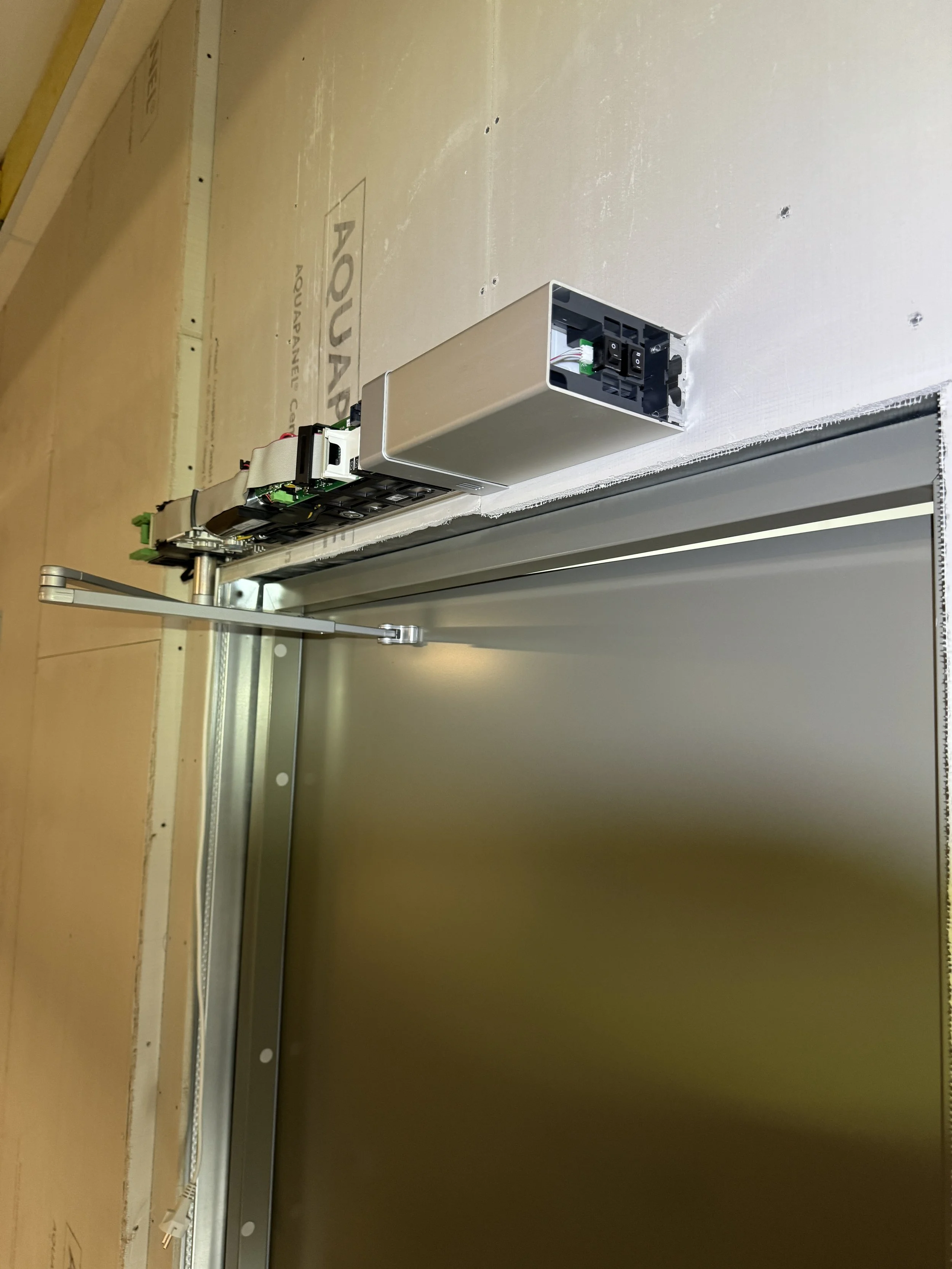 Innendørs konstruksjon med metallskinne og elektroniske komponenter, muligens en automatisert dørmekanisme under installasjon.