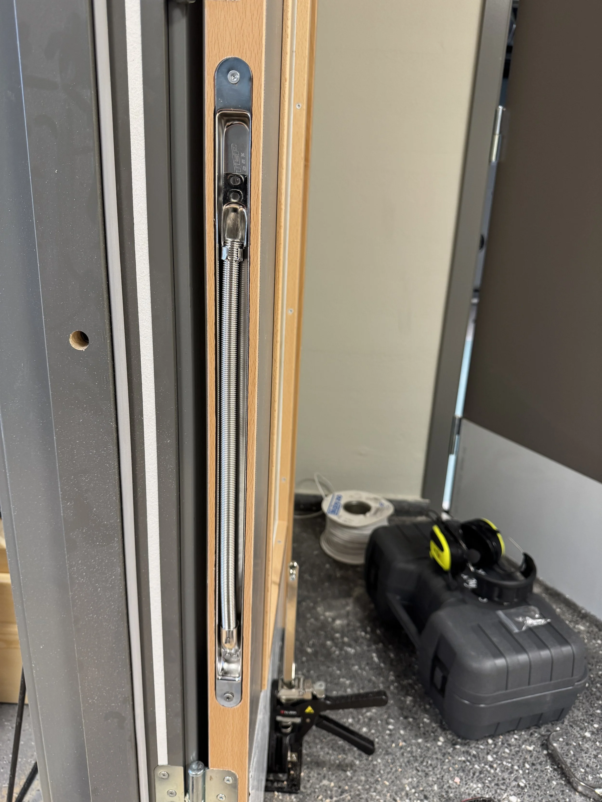 Nærbilde av en døråpning med en metall- og treramme, der det er installert en elektrisk lås eller spærre mekanisme. Det er verktøy og utstyr på gulvet, inkludert en svart koffert, en målebånd, og elektrisk kabel.