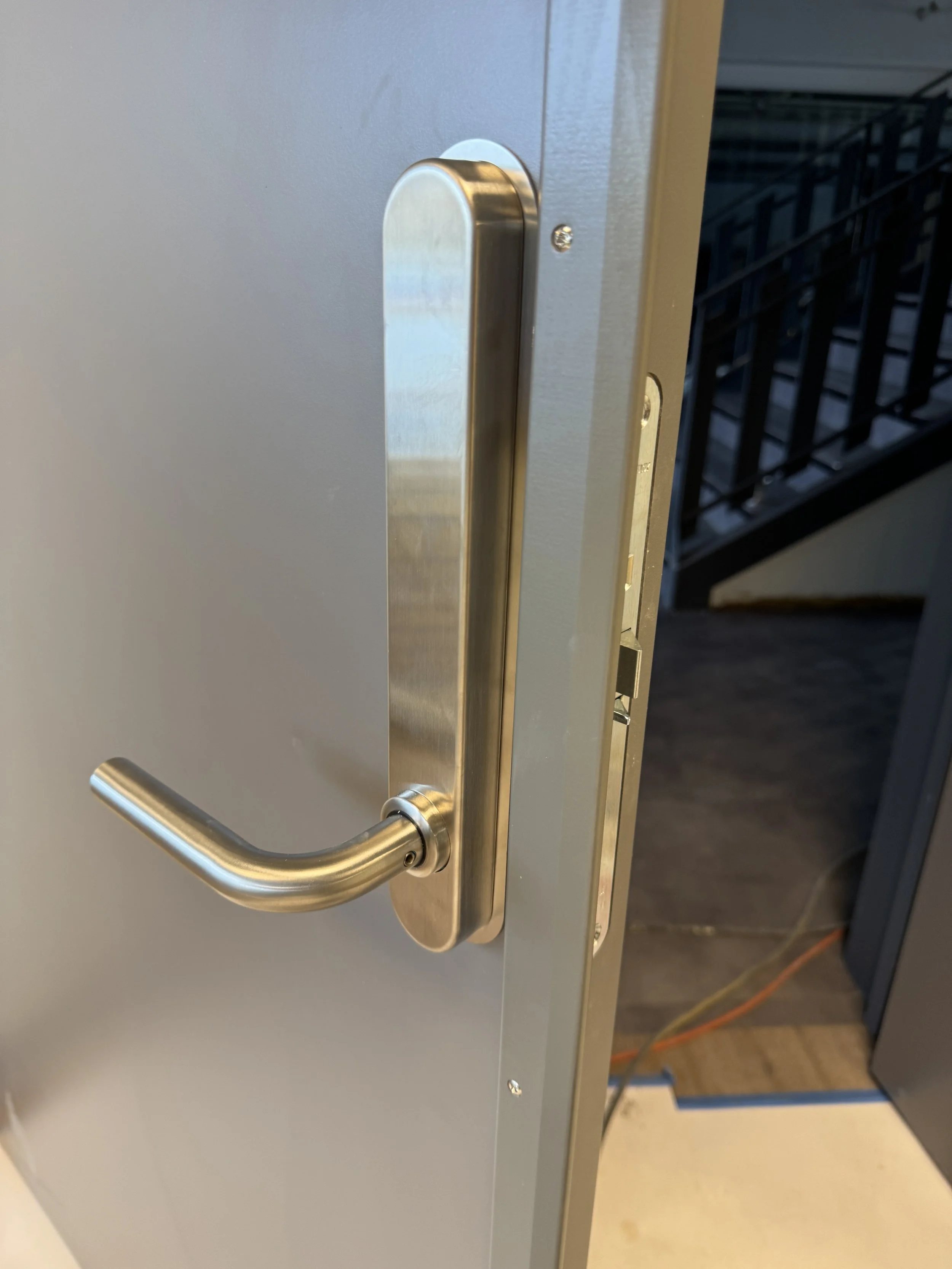 Nærbilde av en metall dørhåndtak på en grå dør, med en synlig låsemekanisme. Bakgrunnen viser trapp og deler av korridor.