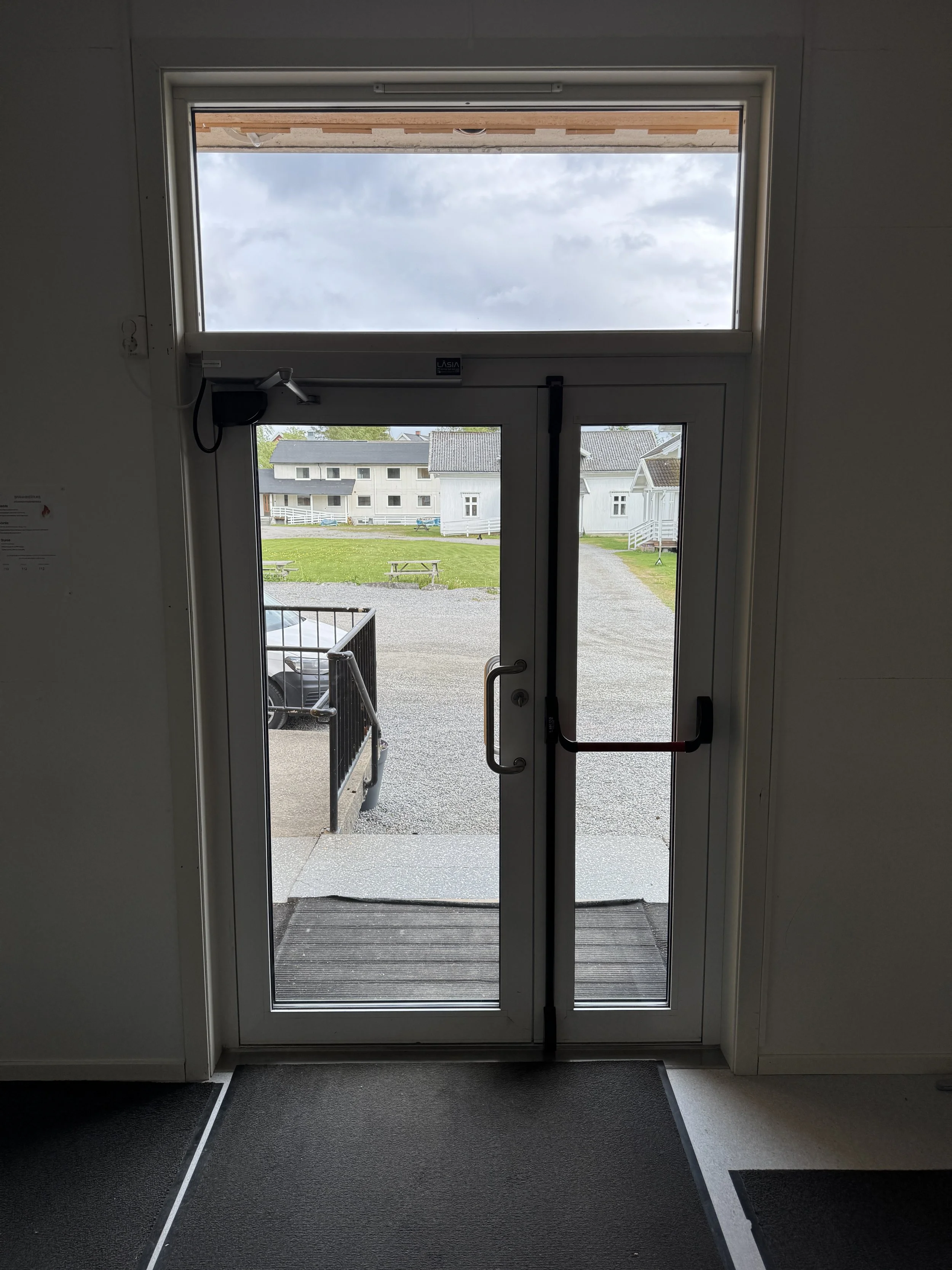 Inngangsdør med vindu og utsikt over parkeringsplass og hage, med overskyet vær