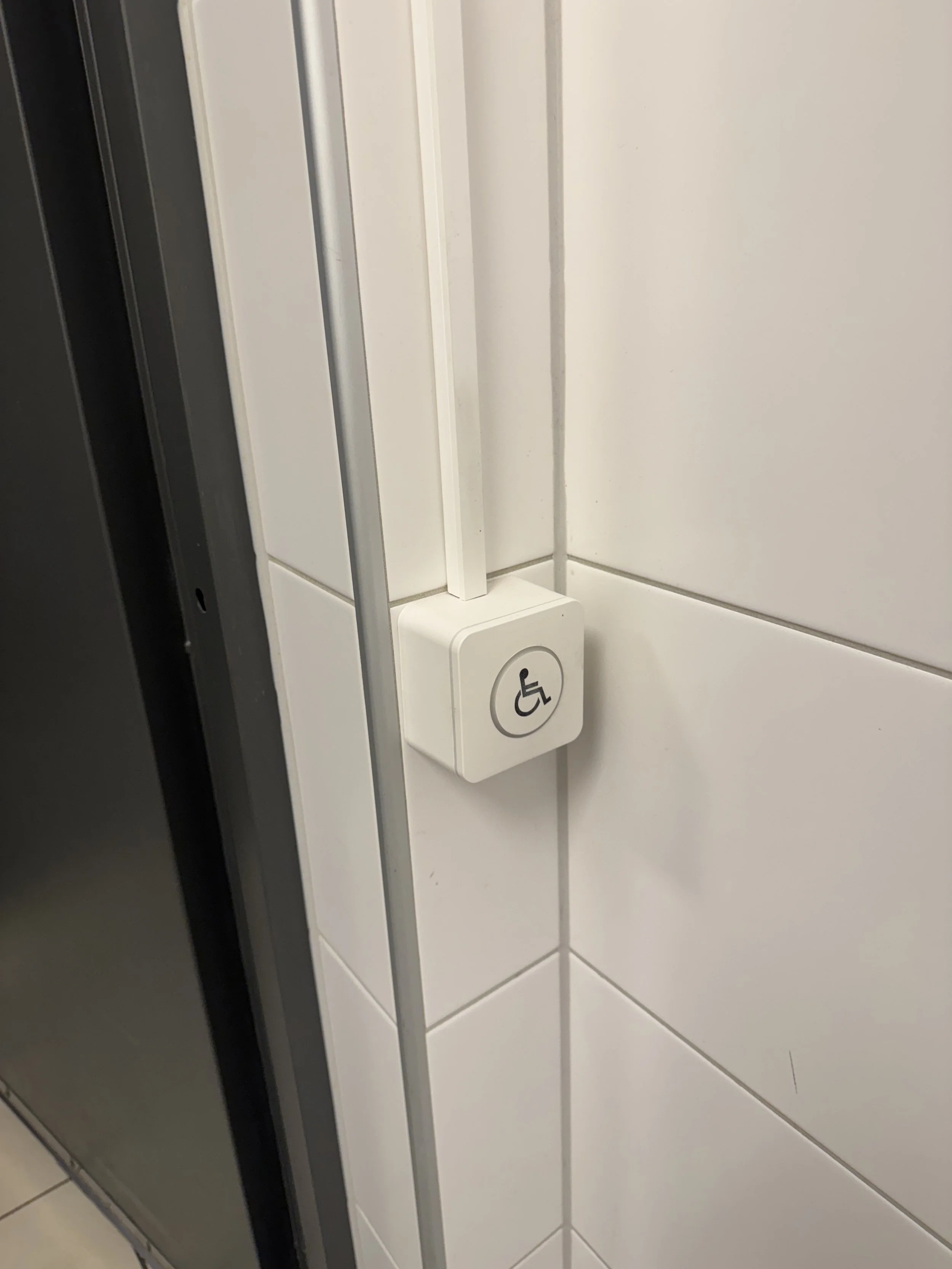 En dør med en knapp for rullestolbrukere, plassert på veggen i et rom med hvite fliser.