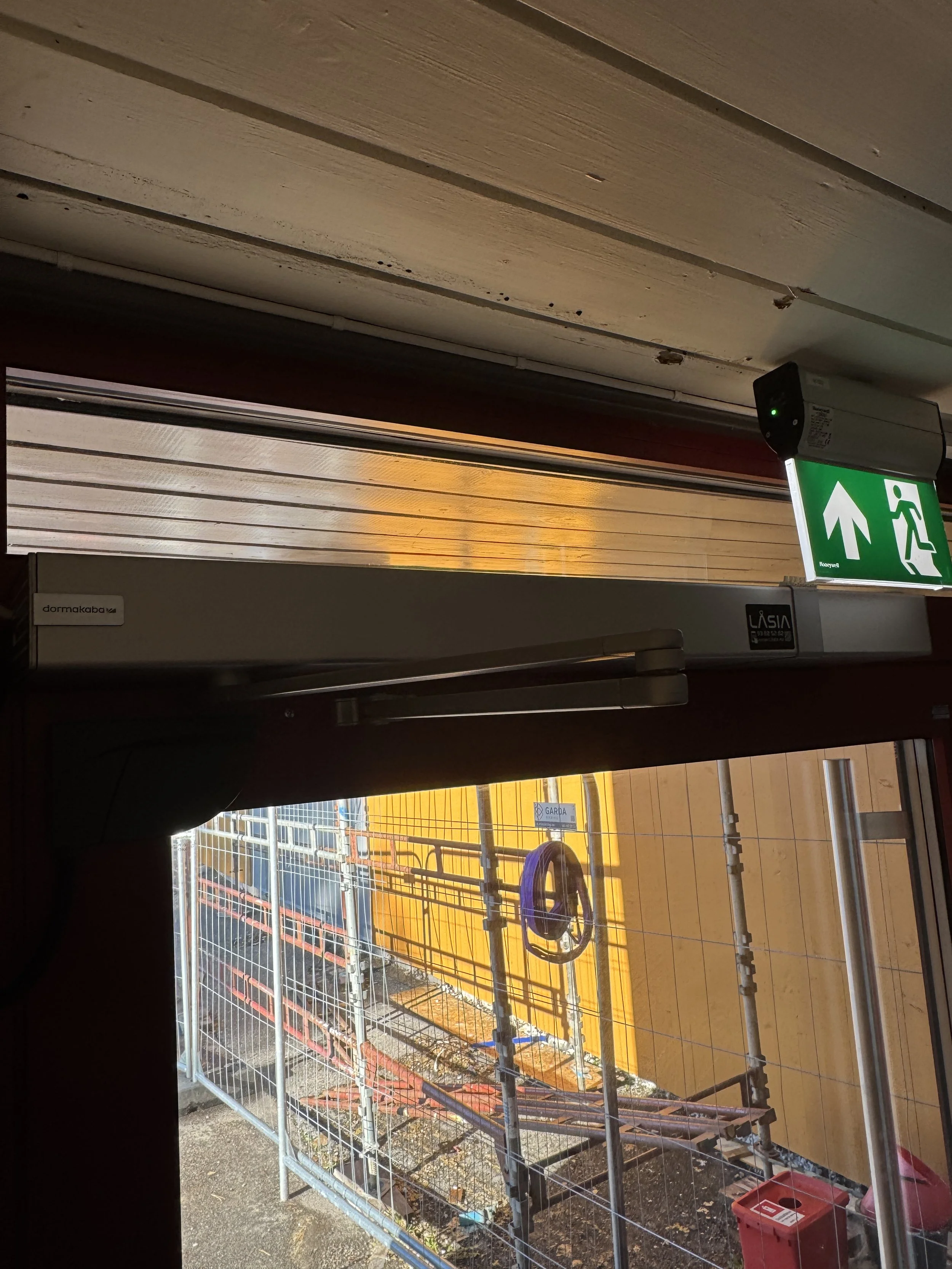 Innendørs bilde av en utgangsdør med en grønne nødutgangssignalet over døren, der den ser ut til å være under bygging eller vedlikehold utendørs.