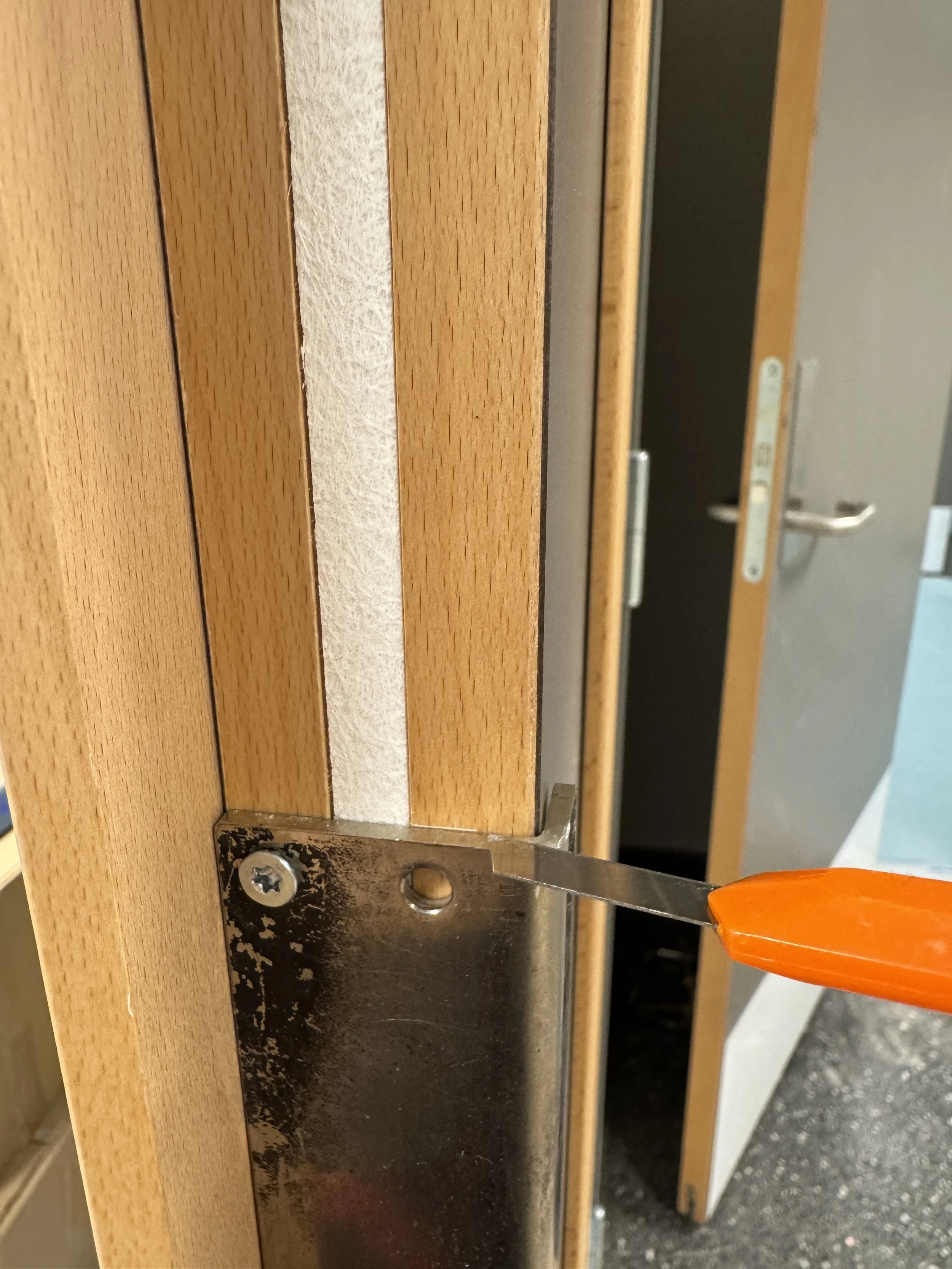 En person bruker en skrutrekker for å skru fast en metallplate til en furukarm. I bakgrunnen er det åpen garderobe med stålstenger og klær.