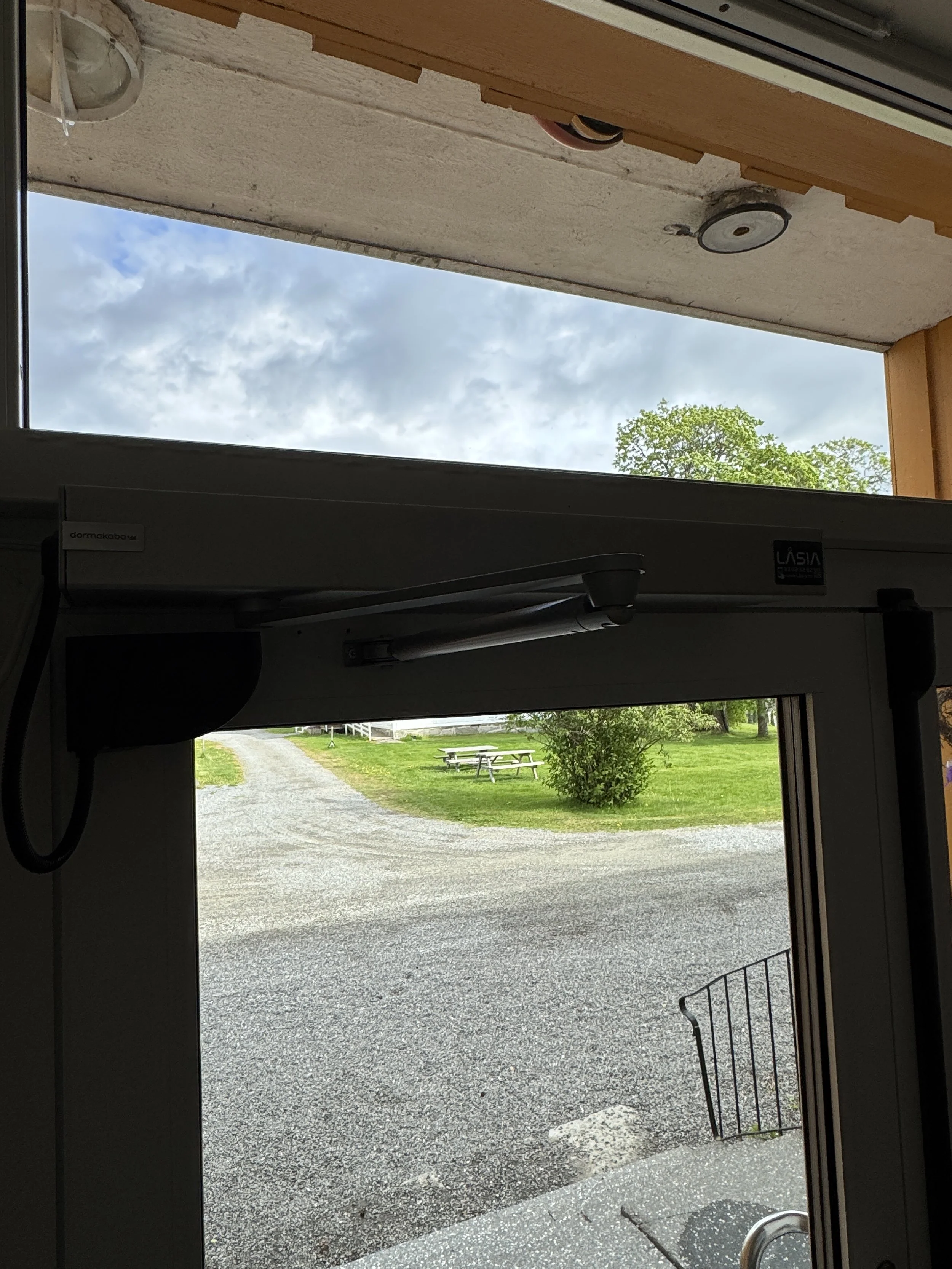 Dette bildet viser utsiden sett gjennom en døråpning, med en grusvei, grønt gress, noen busker og trær, samt benker i landskapet. Over er en del av taket og noen lys. Himmel med skyer.