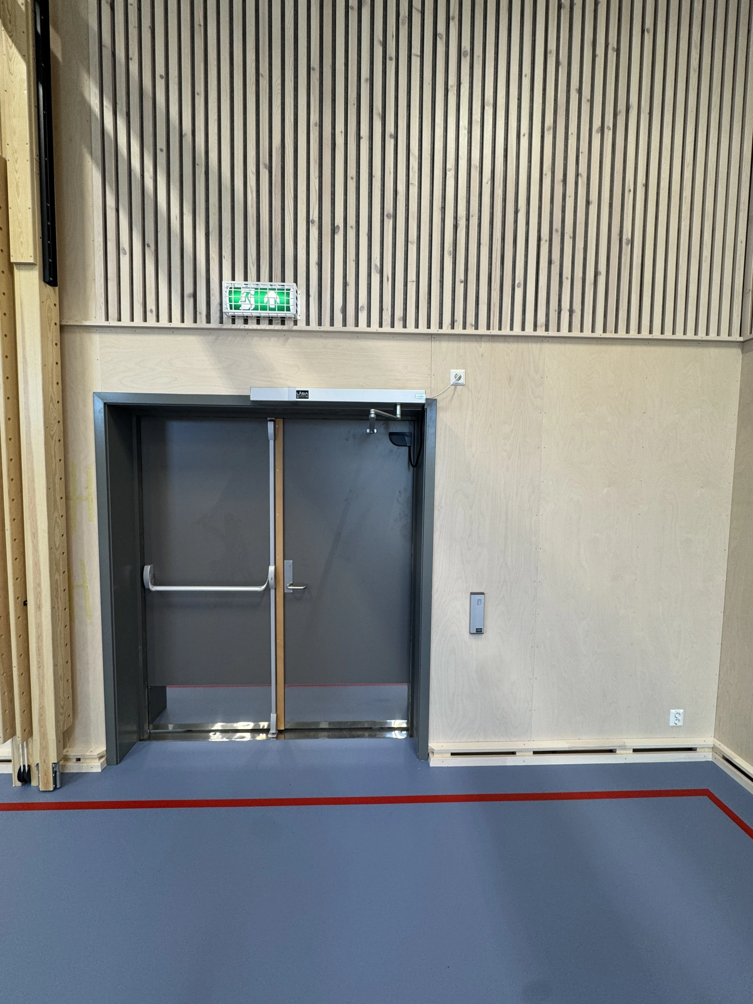 Innendørs bilder av en dør med brannvernskilt og nødutgangssymbol over, i et rom med veggpaneler og en grå gulvflate med rød markering.