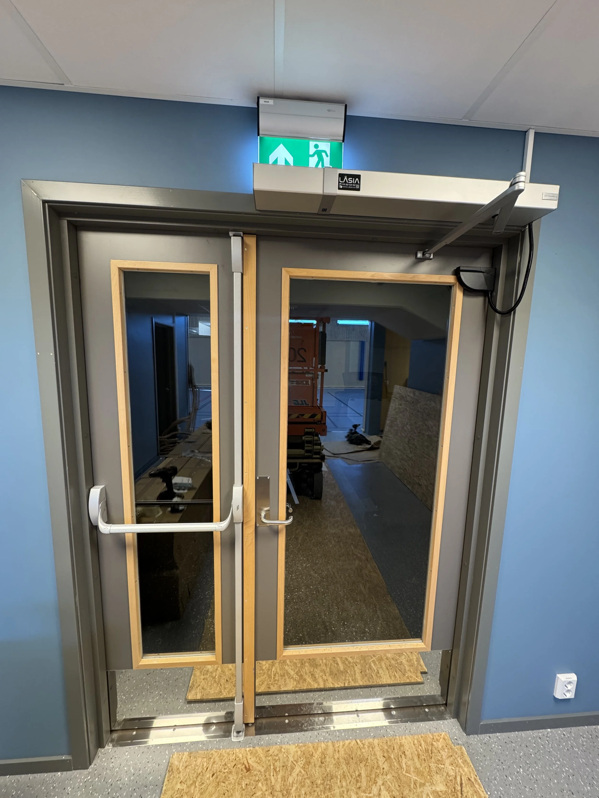 Dørene til en inngang med glassvinduer og en automatisk dørlukker, med en nødutgangsskyvegruppe med lysende grønt evakueringsskilt over.