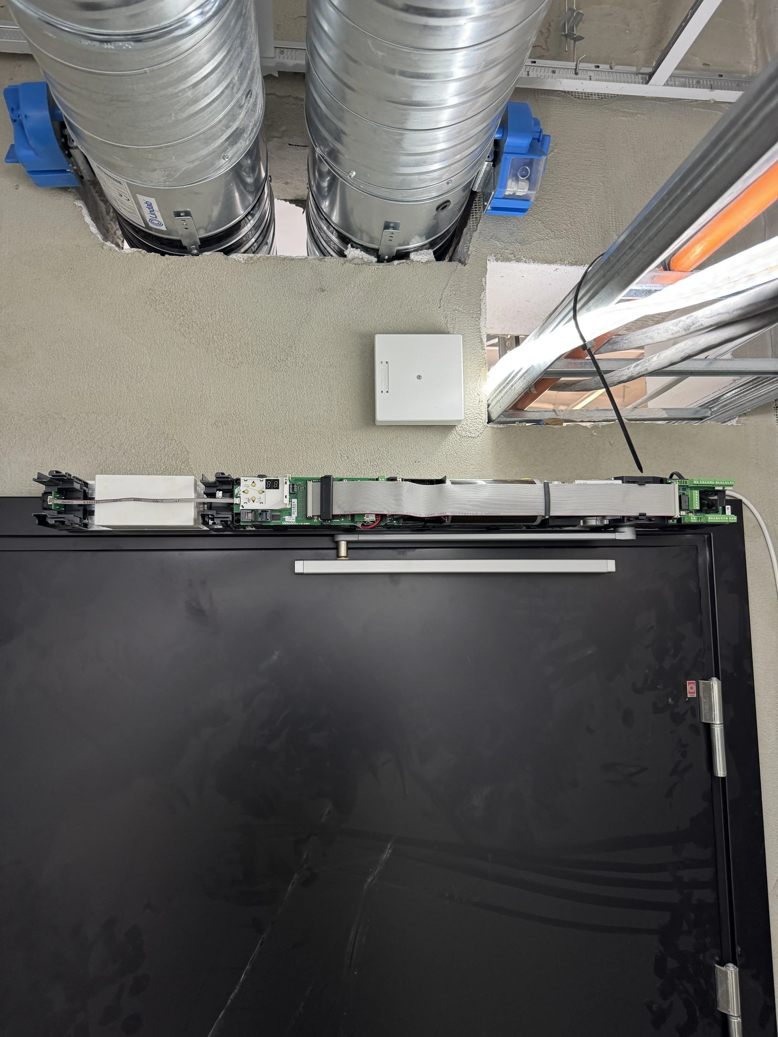 Bildet viser en innendørs teknisk installasjon med metallrør, elektrisk utstyr og en stor svart dør eller panel, sannsynligvis en del av et ventilasjons- eller teknisk rom.