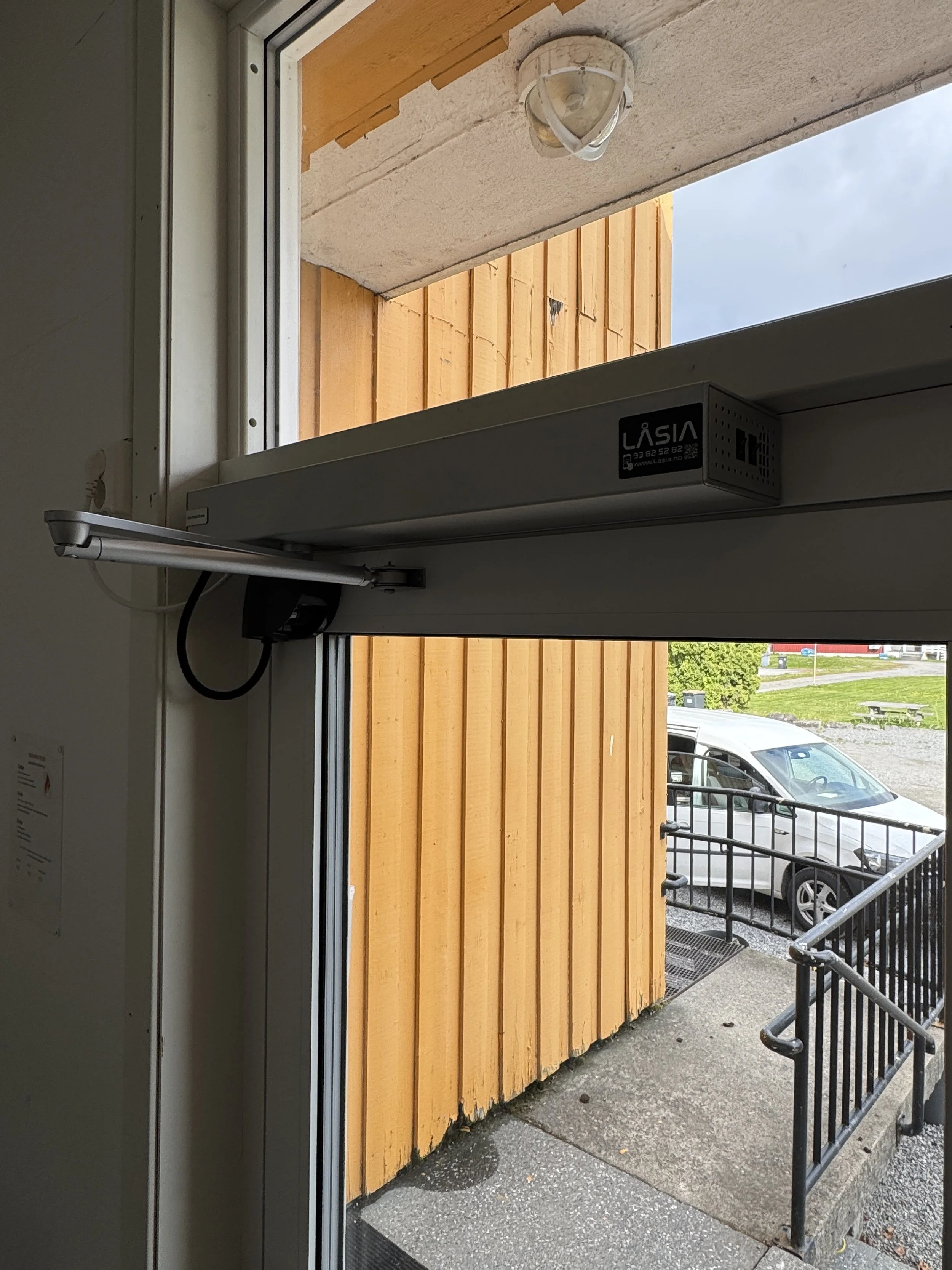 Innvendig med automatisk døråpner, som åpner en utgang mot en liten uteplass med gjerde og bilparkering. Ute er det en bygning i tre med gul maling.