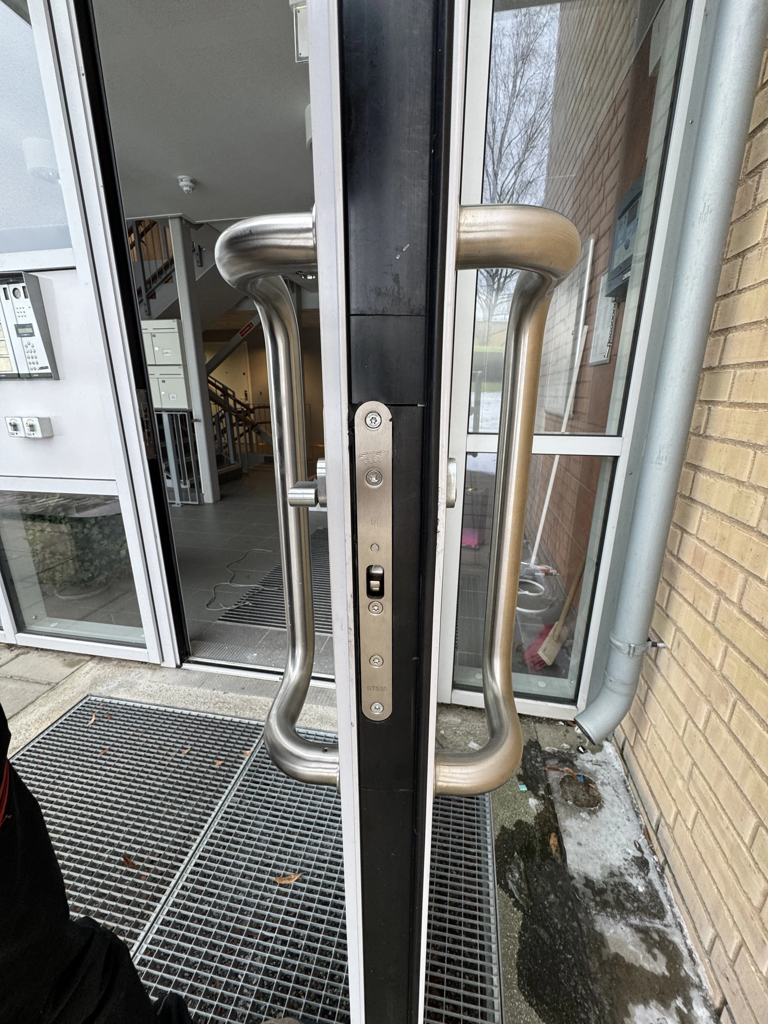 Nærbilde av en glassdør med metallhåndtak uten synlige nøkler, og en bygning med mursteinvegg og et inngangsparti med mattene foran døren.