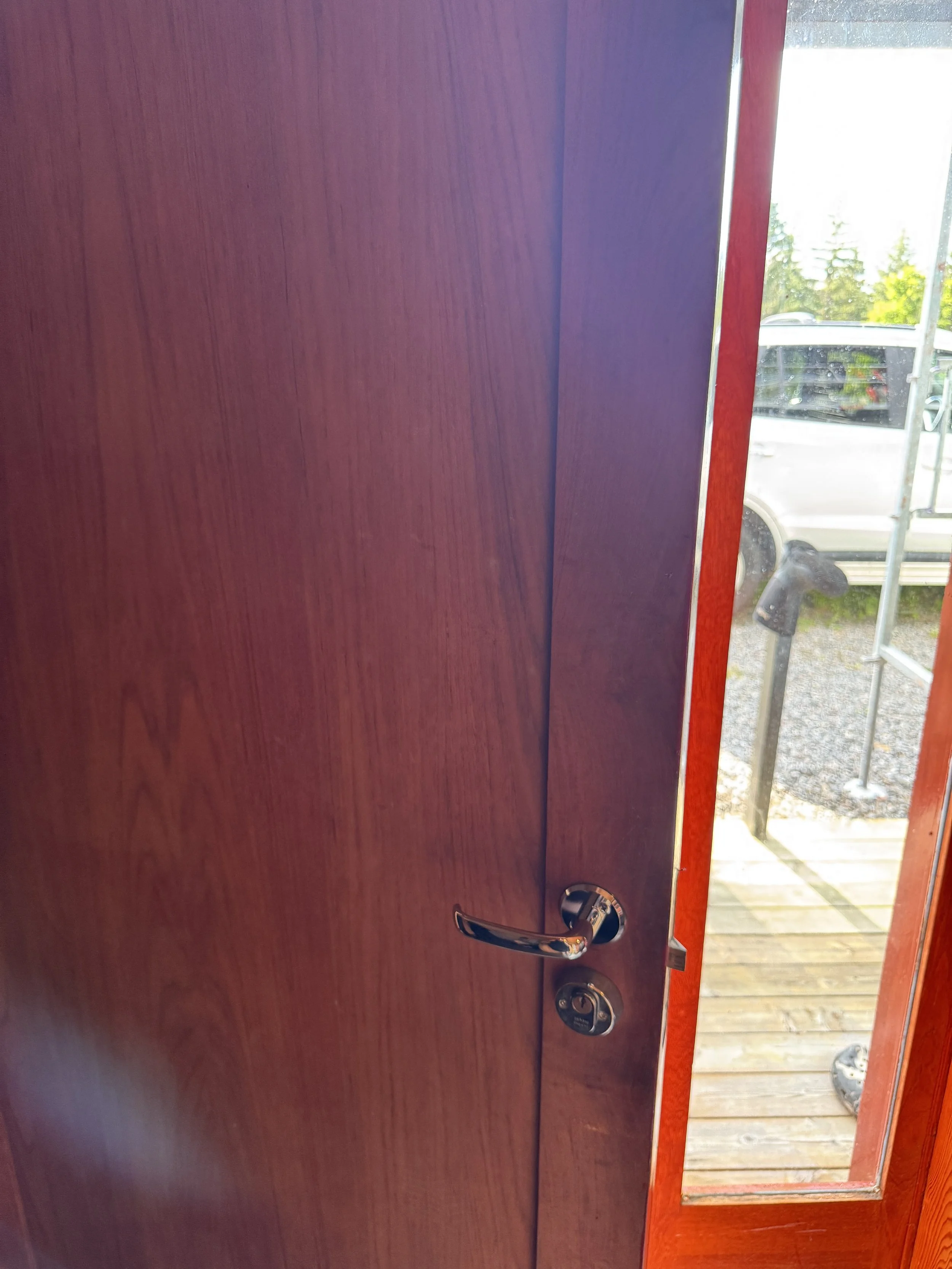 Et tre- eller maltøyet tre- eller kombinasjonsdør med glassfelt på siden, som åpnes ut mot en utendørs terrasse eller gangvei med trapp og utendørsutstyr.