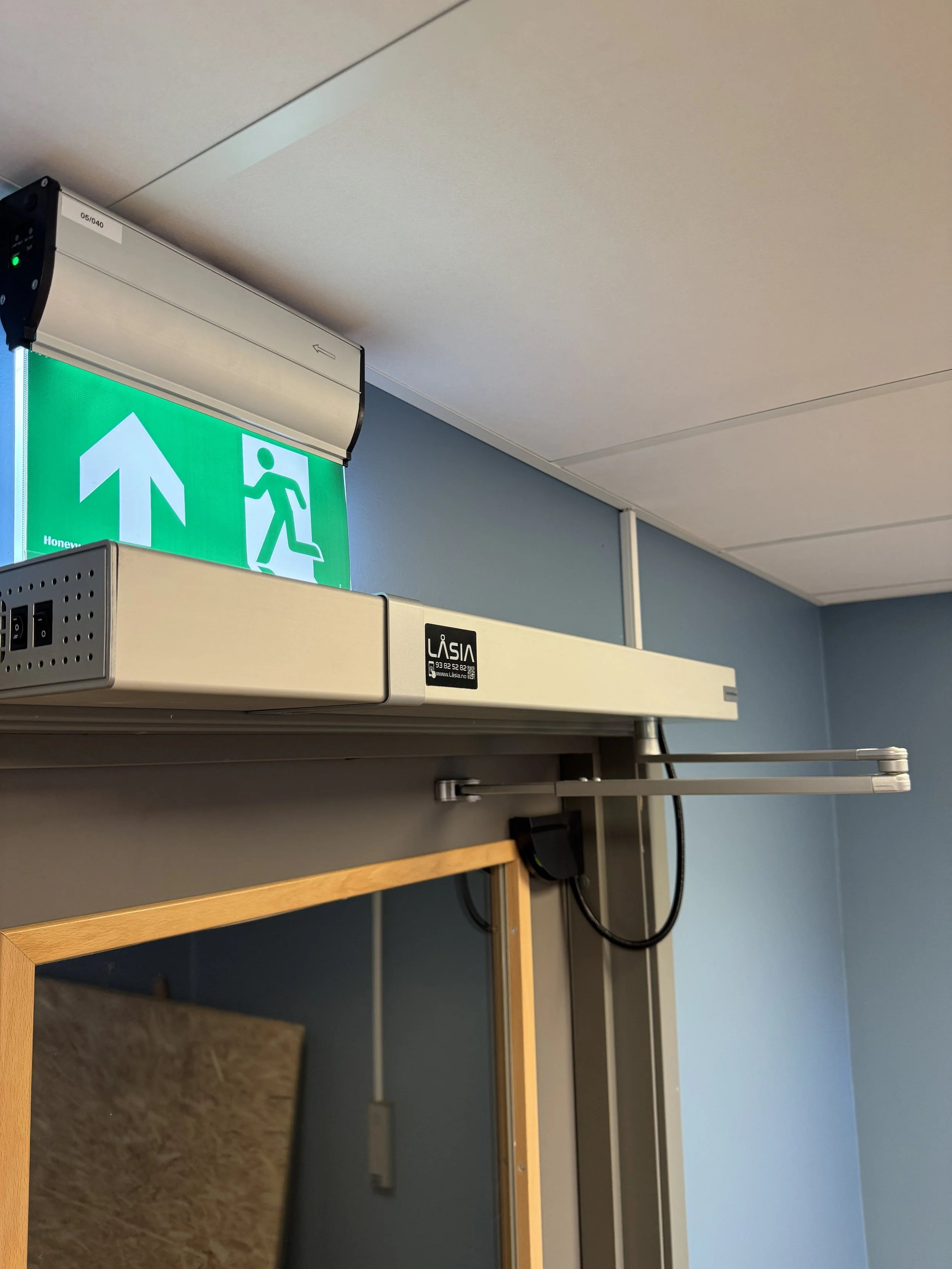 Nærbilde av en nødutgangssignatur med grønn bakgrunn og hvit pil som peker oppover, plassert over en dør i et rom med blåvegger.