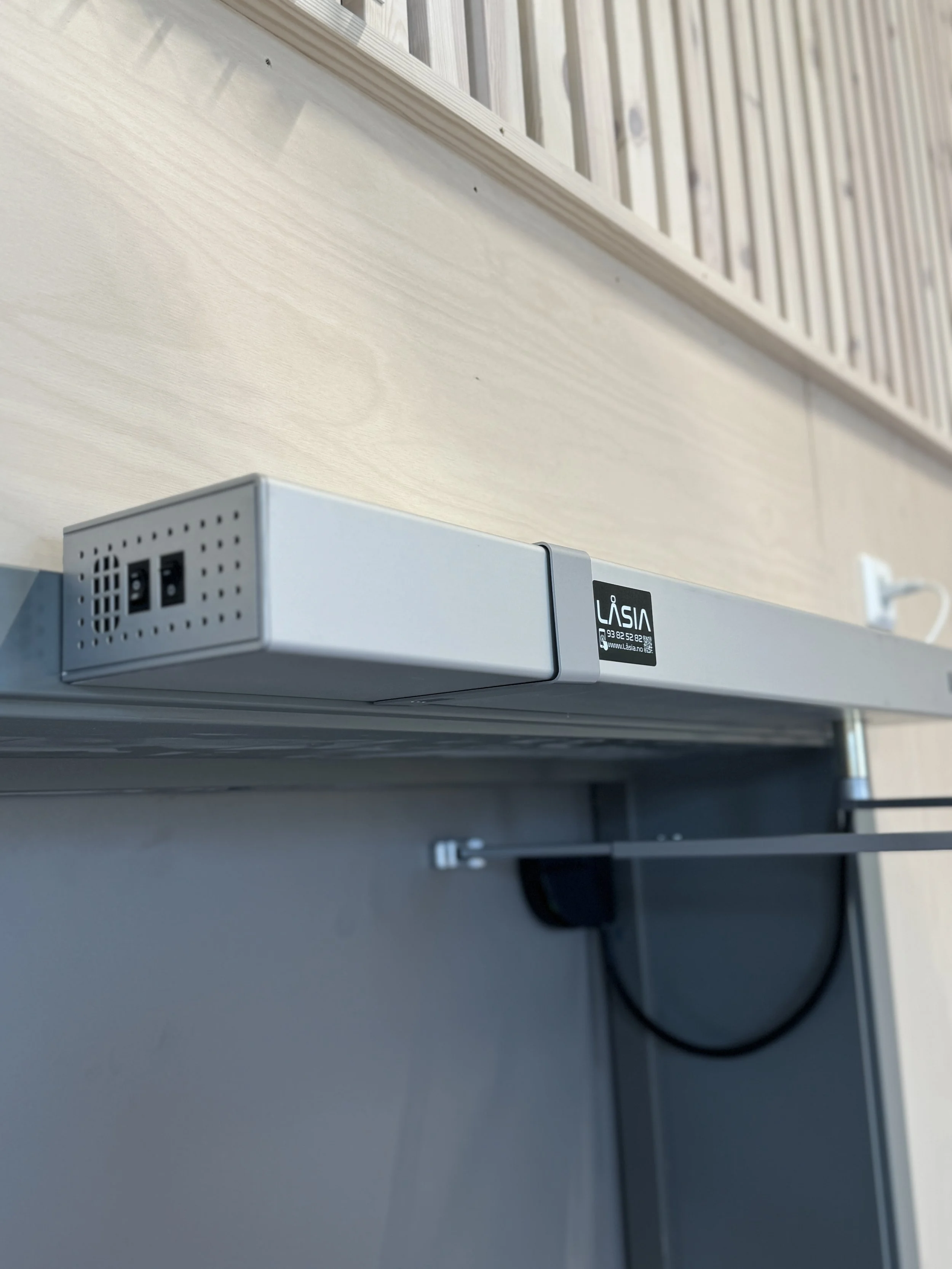 En elektrisk LED-stripe montert under en lys trevegghylle, med en liten merkelapp som sier LASIA.