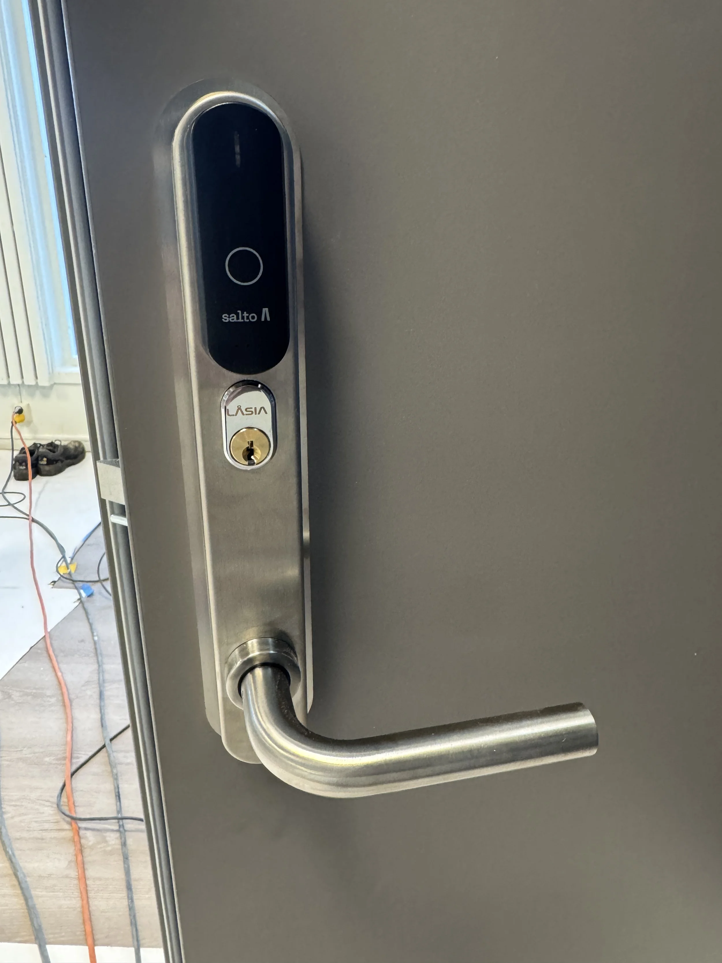 Nærbilde av en dørhåndtak med elektronisk adgangskontrollsystem, som har en svart sensor samt en nøkkellås med merket LASIA.