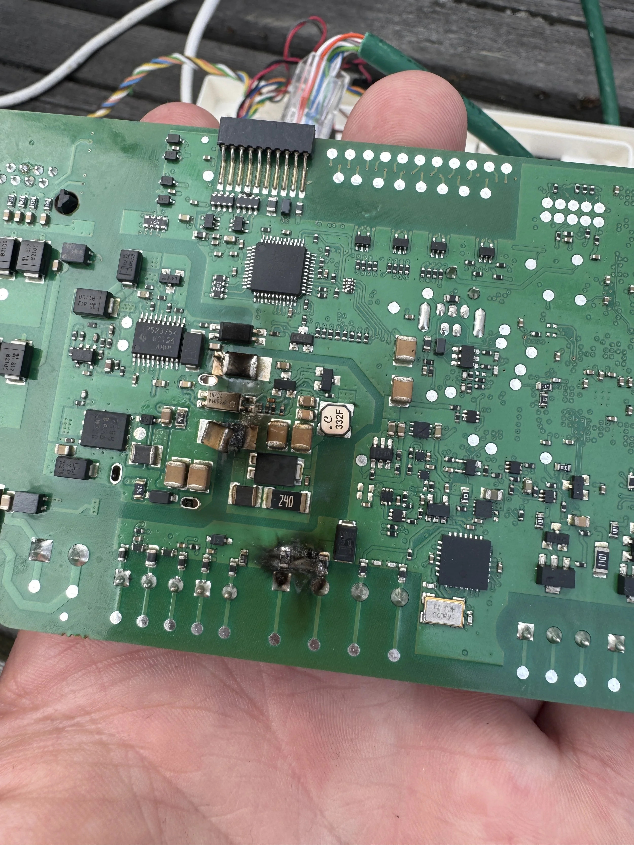 En person holder en brent kretskort med brente områder og svarte brannskader, omgitt av flere tilkoblede ledninger.