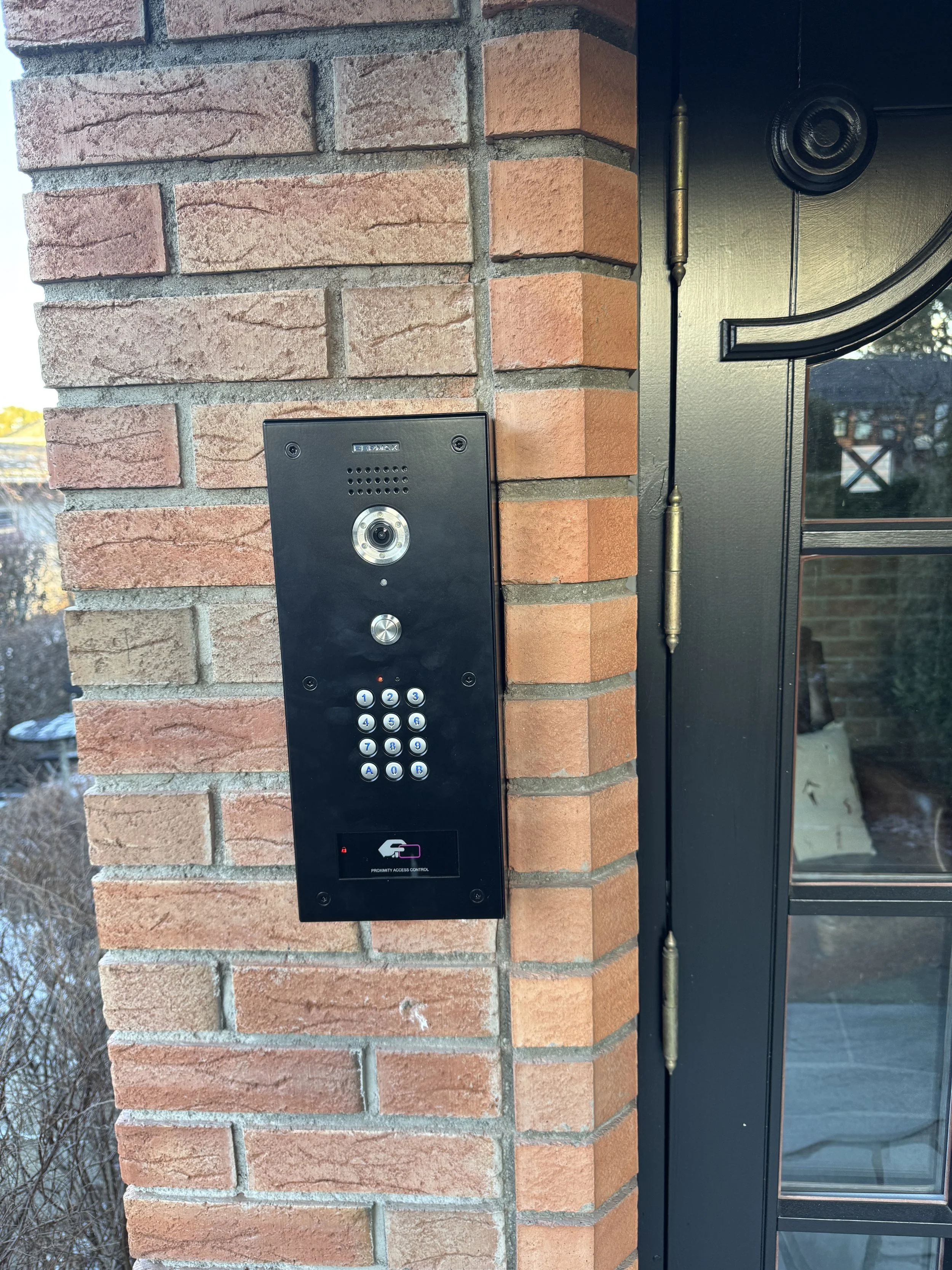 Nærbilde av en sort dørtelefon med tastatur og kamera montert på en murvegg utenfor en inngangsdør.