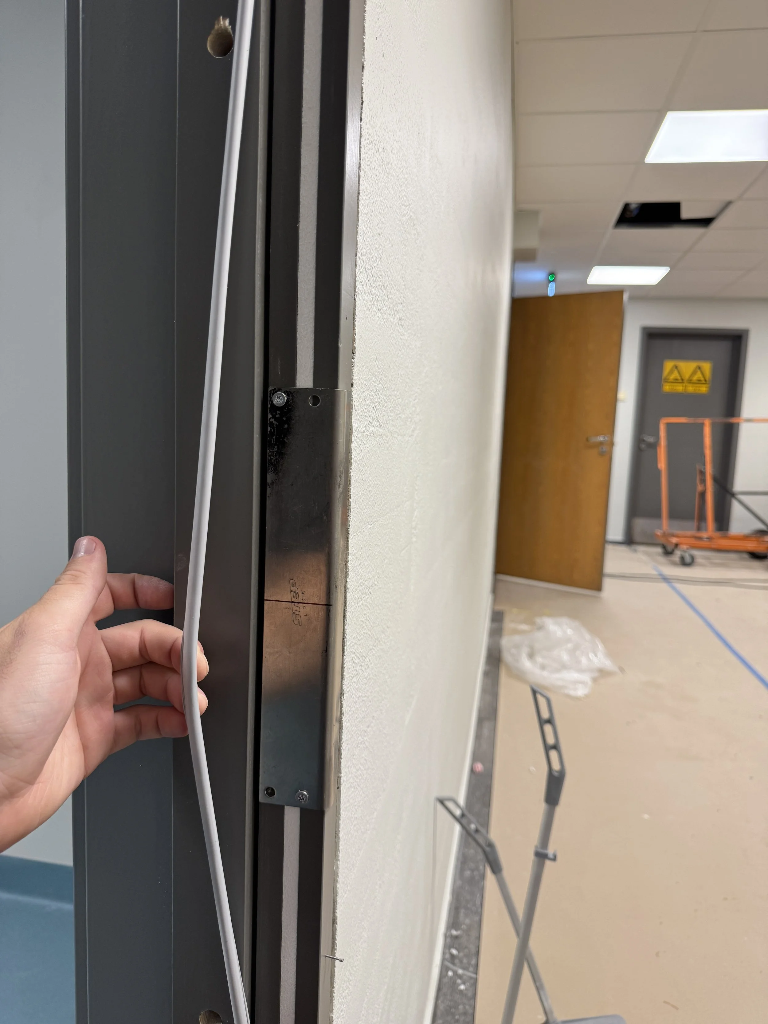 Person som holder et metallramme foran en vegg i en bygning under bygging eller renovering, med en åpen dør og byggeutstyr i bakgrunnen.