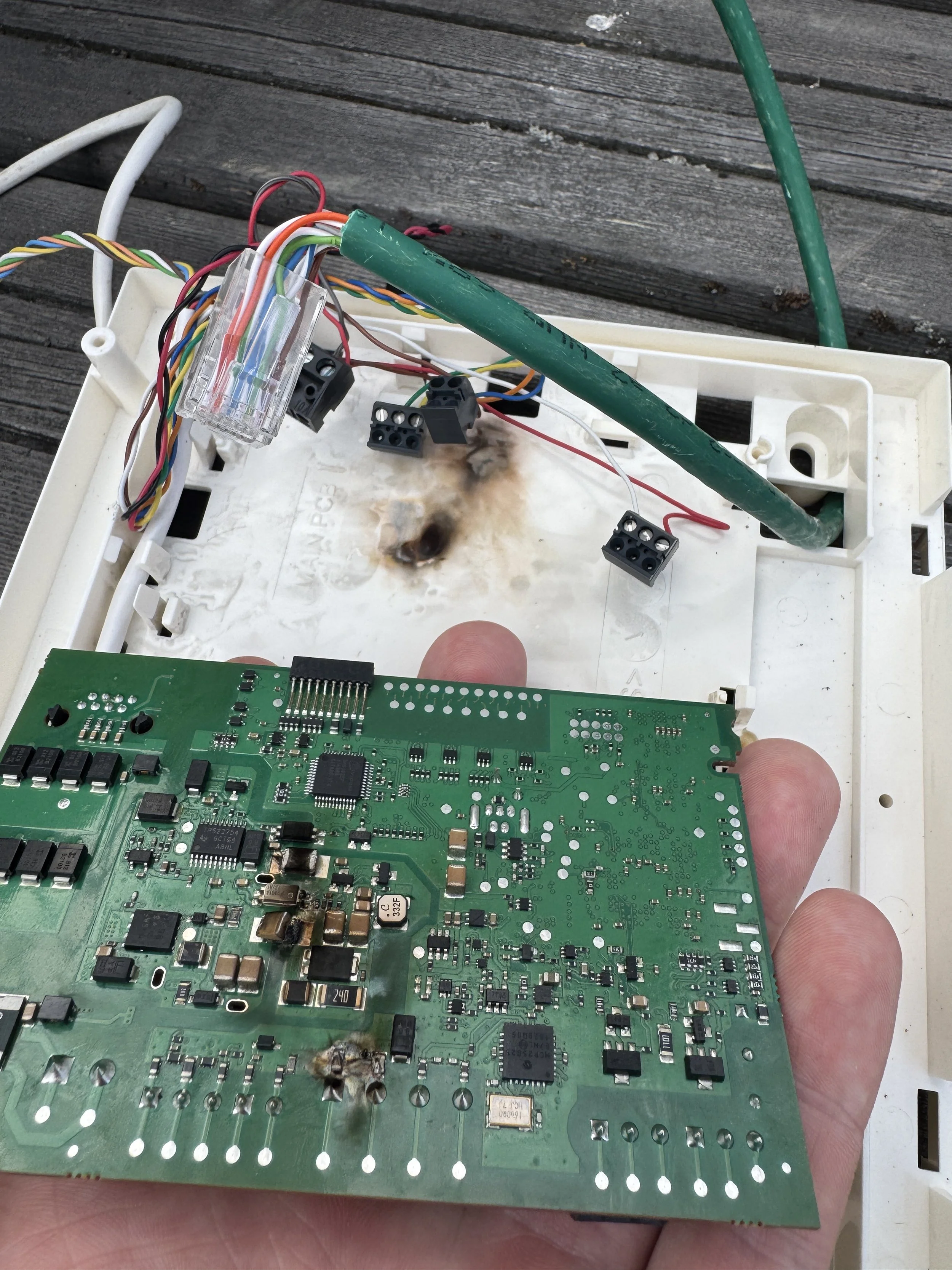 Et ødelagt elektronikkort med brente områder, inne i en hvit plastkasse med flere løse ledninger og kontakter, plassert på en utendørs tre overflate.