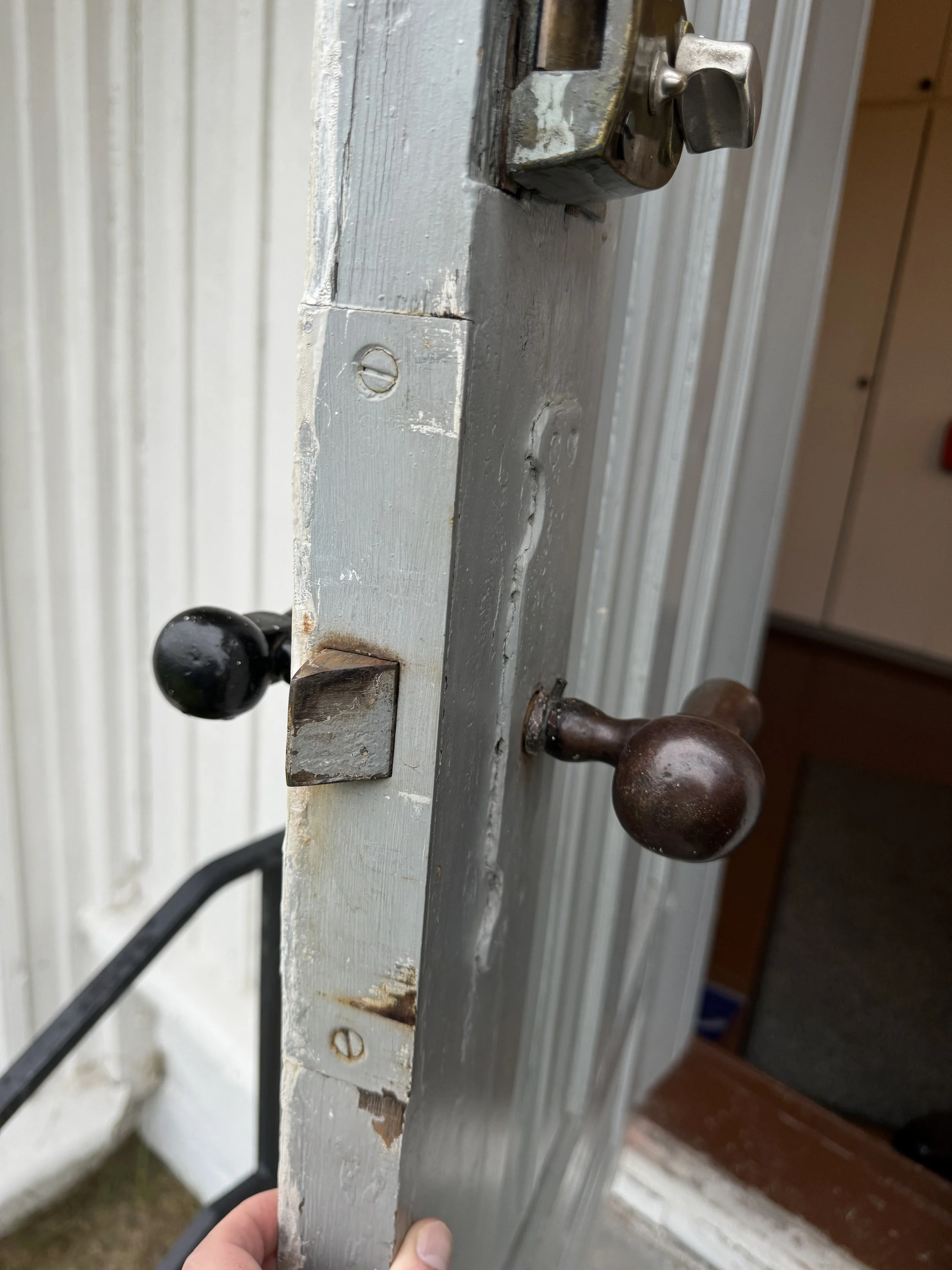 Nærbilde av en eldre, værbitt ytterdør med tre håndtak, ett i svart og to i rustent brunt, festet til en gråmalte treboksdør.