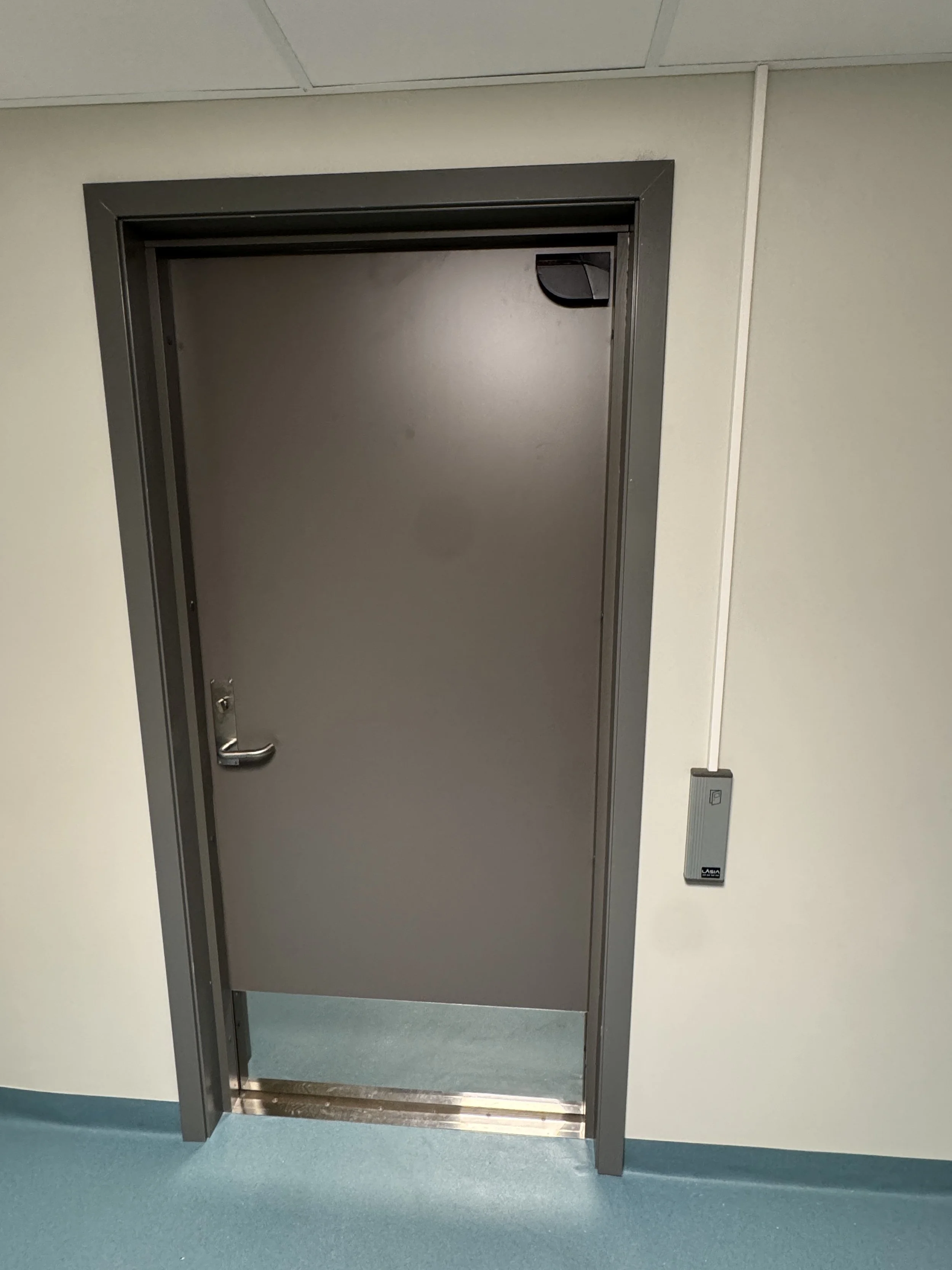 En grå dør i en vegg med en håndtak og en kontrollboks ved siden av, plassert i et rom med lyseblå gulv.