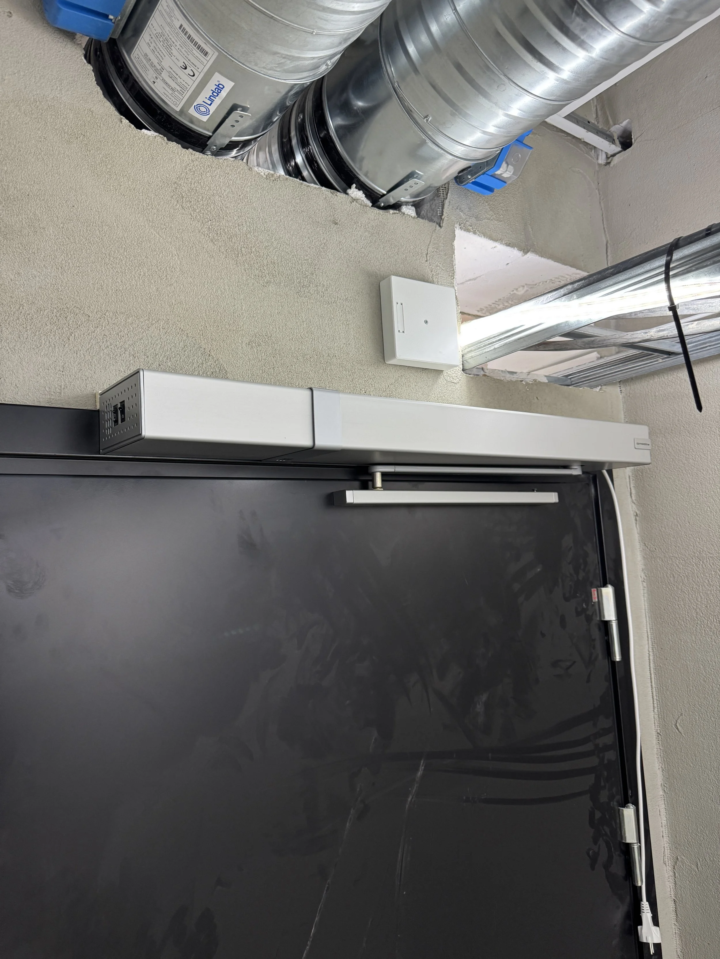 Nærbilde av ventilasjonssystem og en montert adgangskontrollsystem over en svart dør i et bygg.
