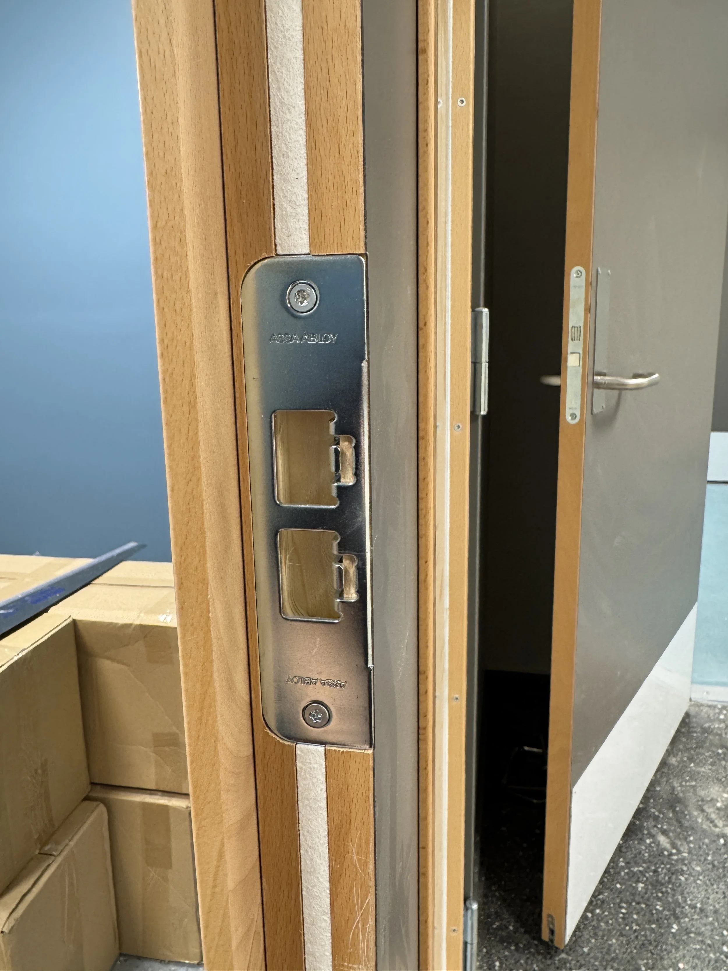 Bildet viser en dørkasse med en metallspak som har to utganger eller tilkoblinger, og en åpen dør i bakgrunnen.