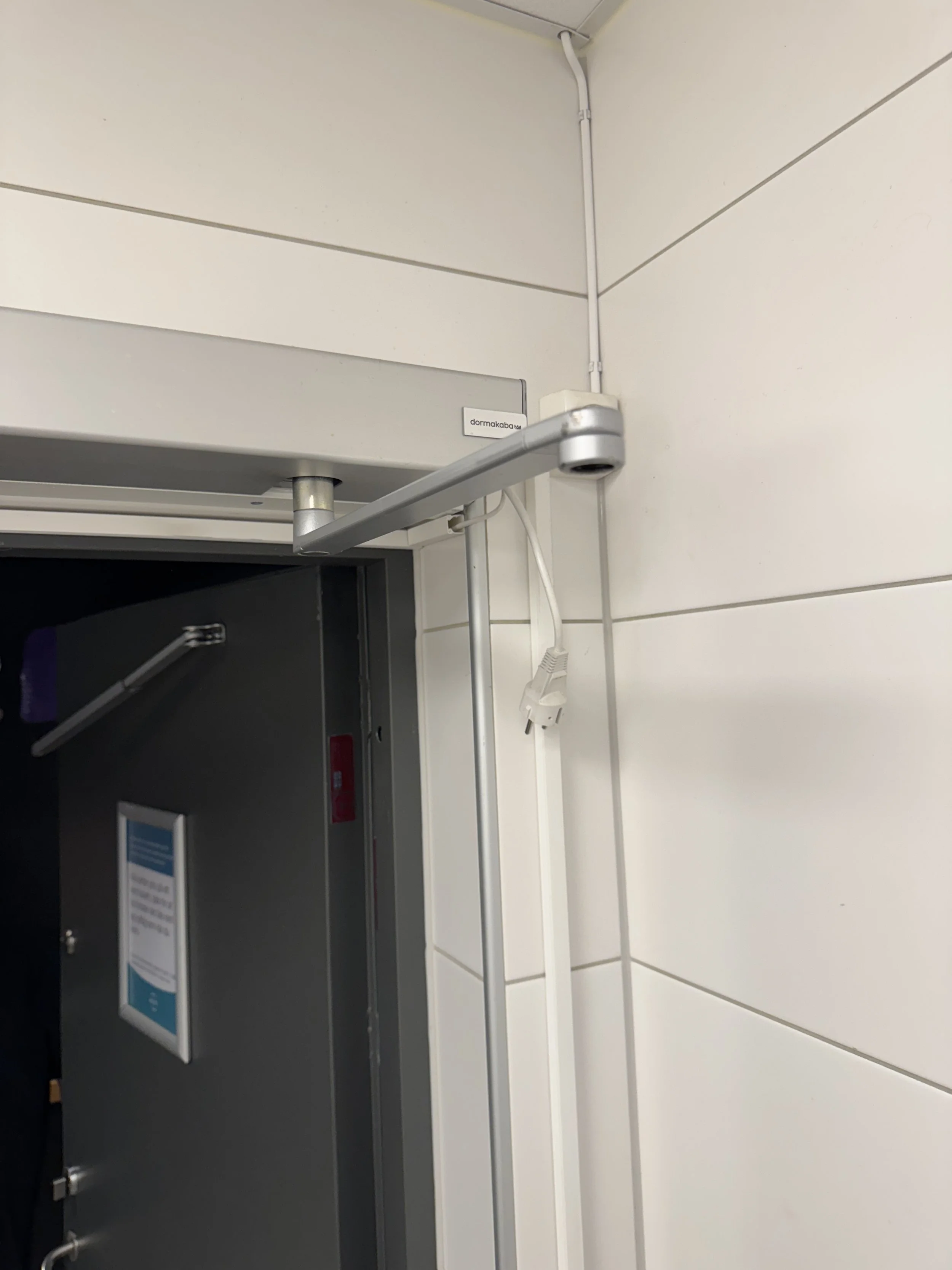 Et hjørne av et rom med hvit vegg og en metallramme, en synlig kabel og en låsbar safe.