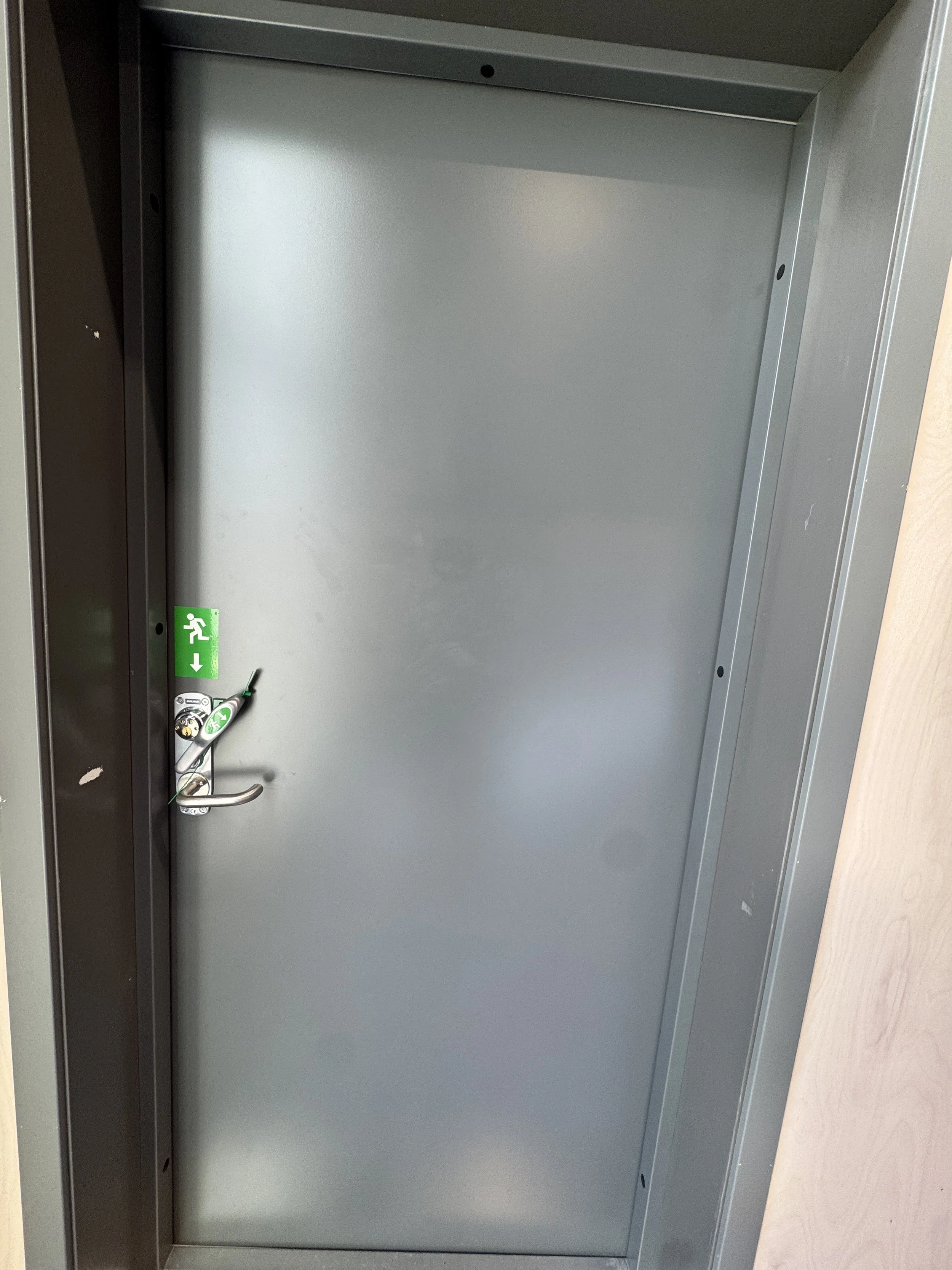 Bildet viser en ståldør med lås og et grønt nødutgangssymbol på venstre side.