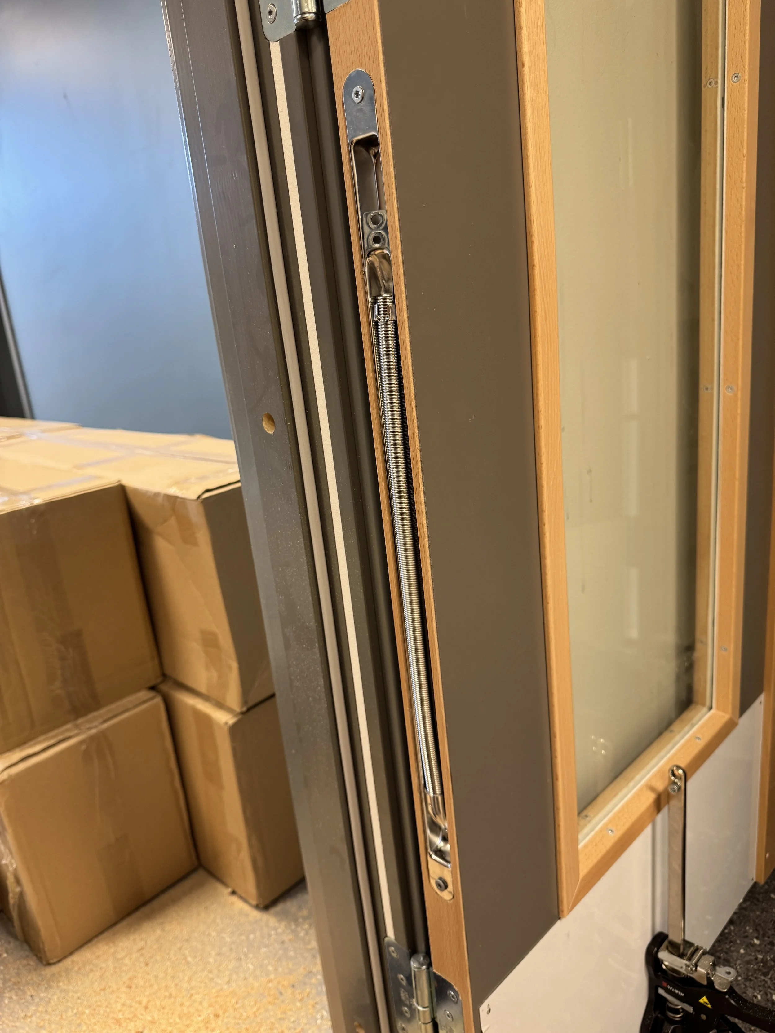 Nærbilde av en mekanisk del på en dør eller et vindu, med en fjær og metallkomponenter.