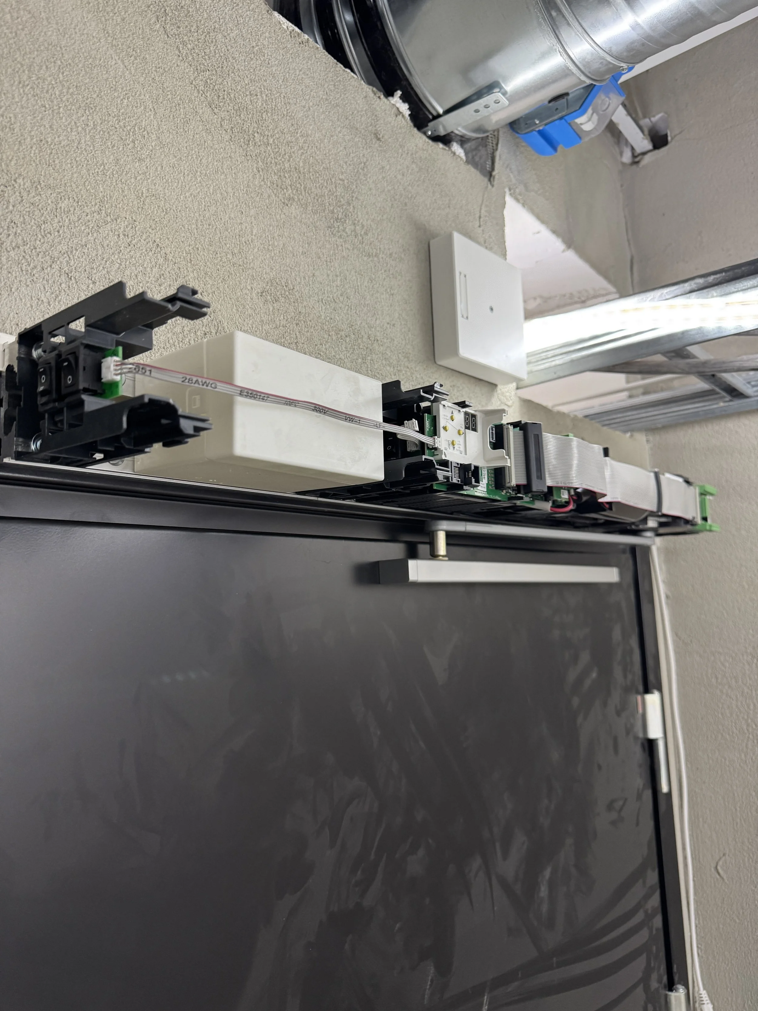 Innendørs bilde av en TV-skjerm som er montert, med synlige elektriske komponenter og tilkoblinger. Bakgrunn viser et vindu med åpen blikk og noe industrielt utstyr.