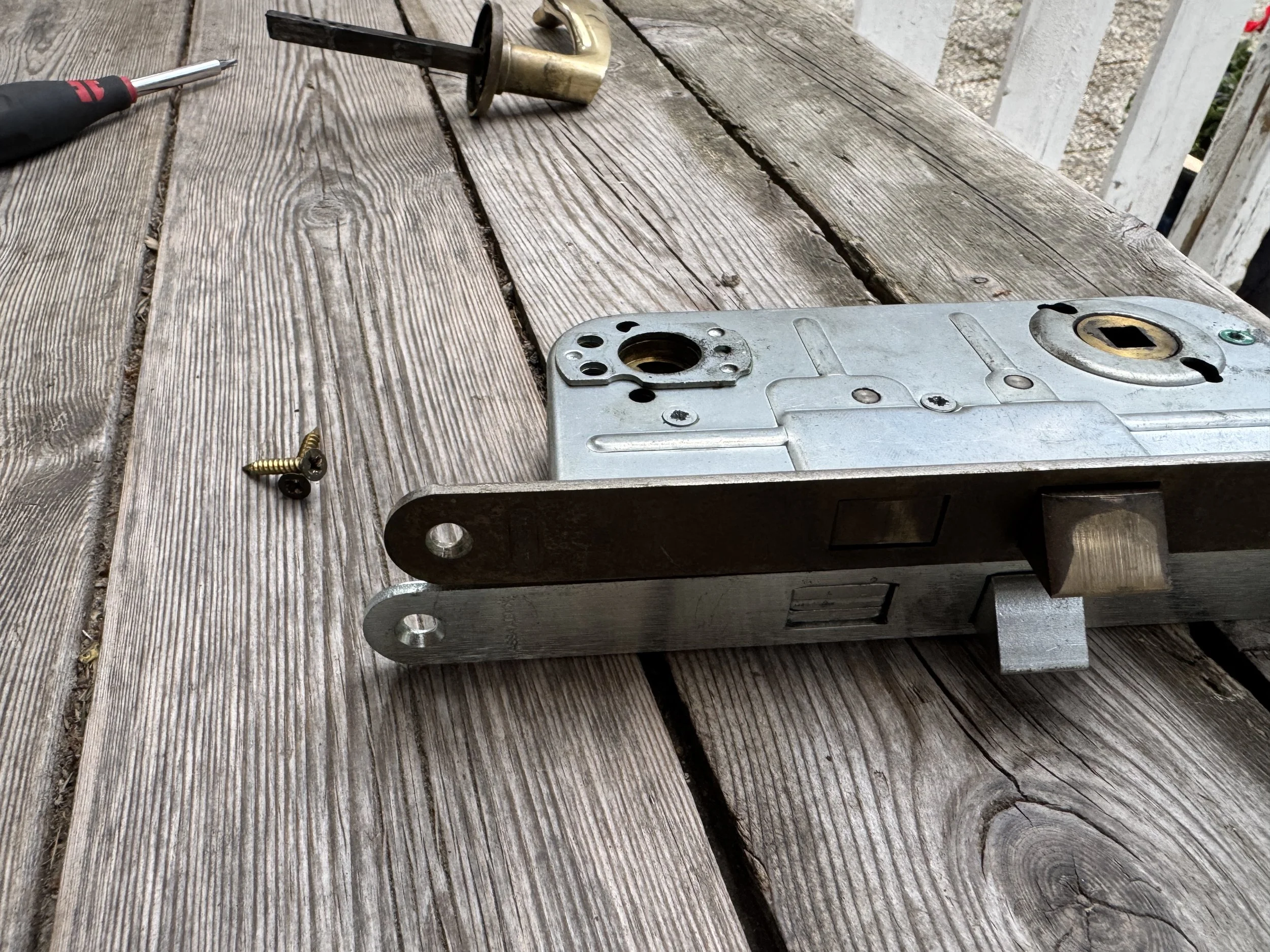 Metal lås med to innretninger, to skruer, en skrutrekker, en skiftenøkkel og et utendørs trebord.