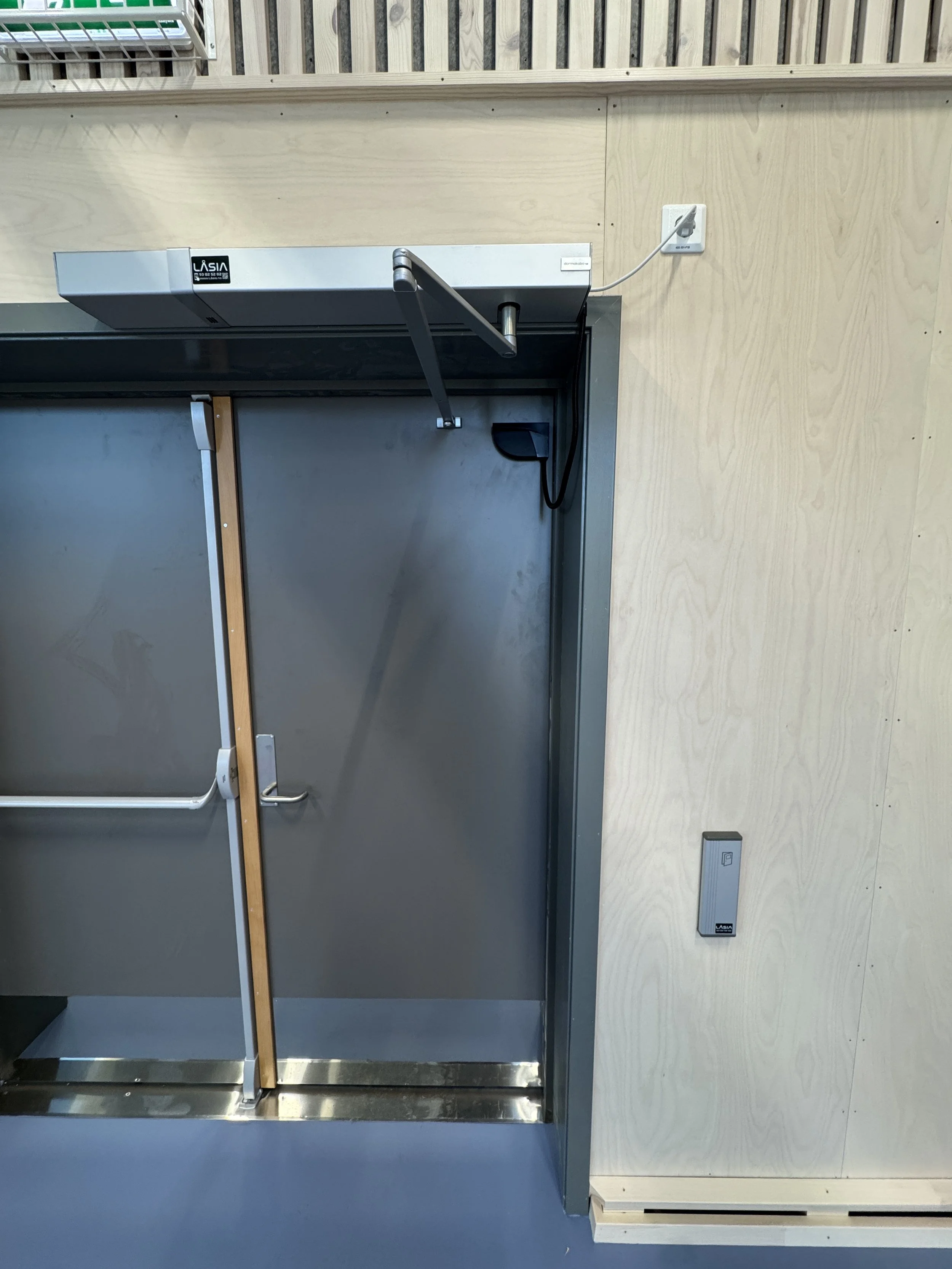 Automatisk døråpner montert over en stålforseglad dør, tilkoblet til en strømkilde. Vegg med lys trepanel og en kort utvendig bryter.