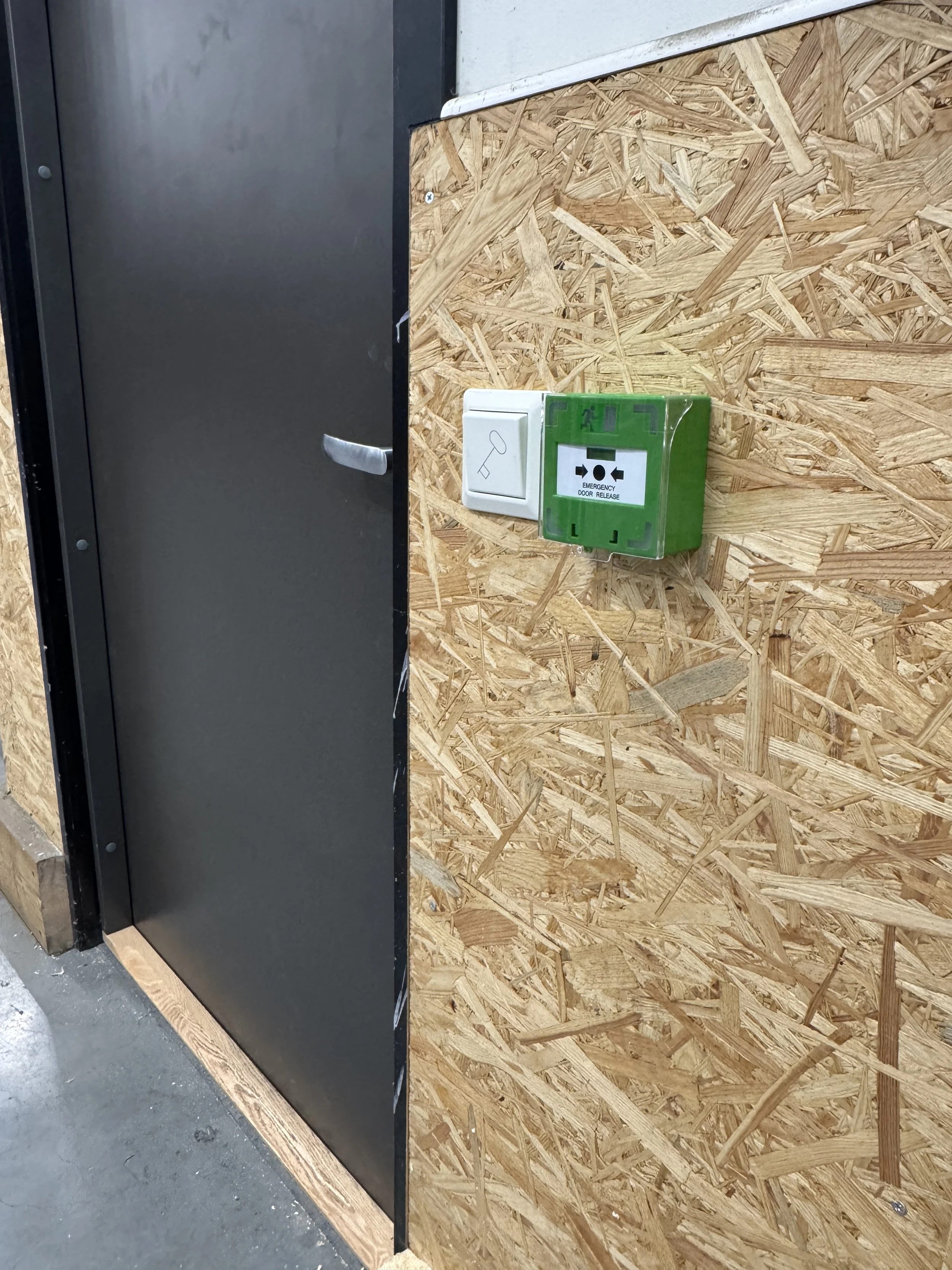 Nærbilde av en vegg med OSB-plater med en hvit knapp og en grønn nødutløserboks for døråpning.