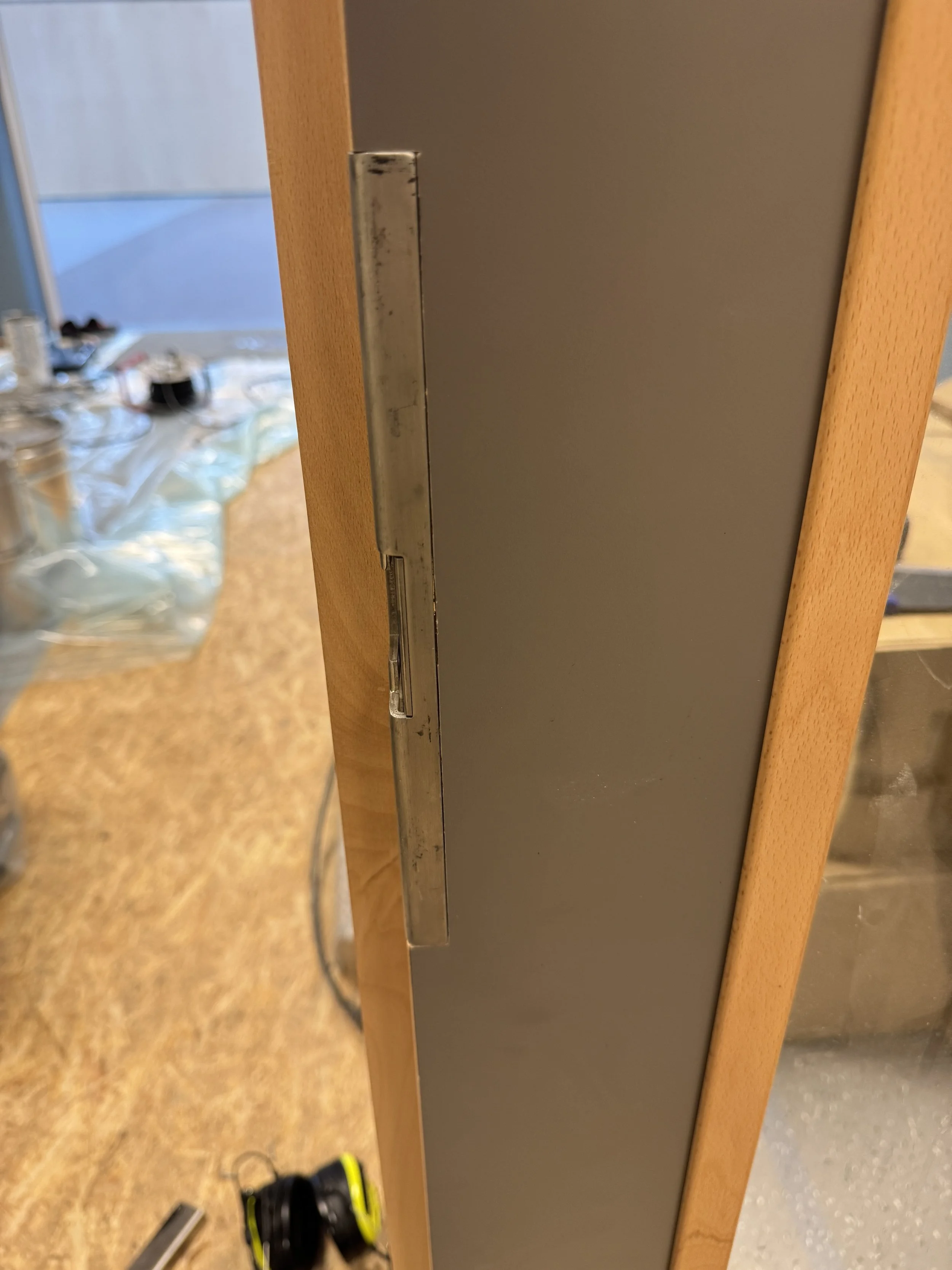 Et bilde av en del av en dørkarm eller et møbel, med en metalldel som kan være en lås eller en håndtakssprosse, på en treramme. Bakgrunnen viser et rom med ulike ting, blant annet verktøy og en arbeidsflate.