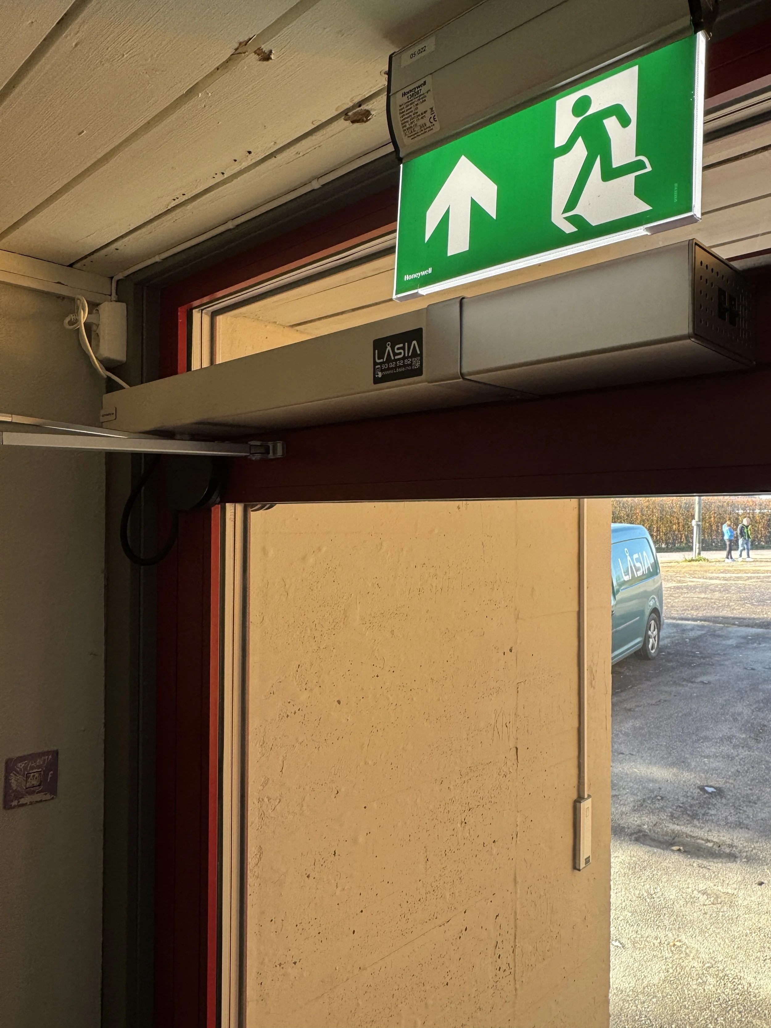 Et grønne utendørs nødutgangsskilt som peker oppover med en løpende person symbol, over en dørvindu.