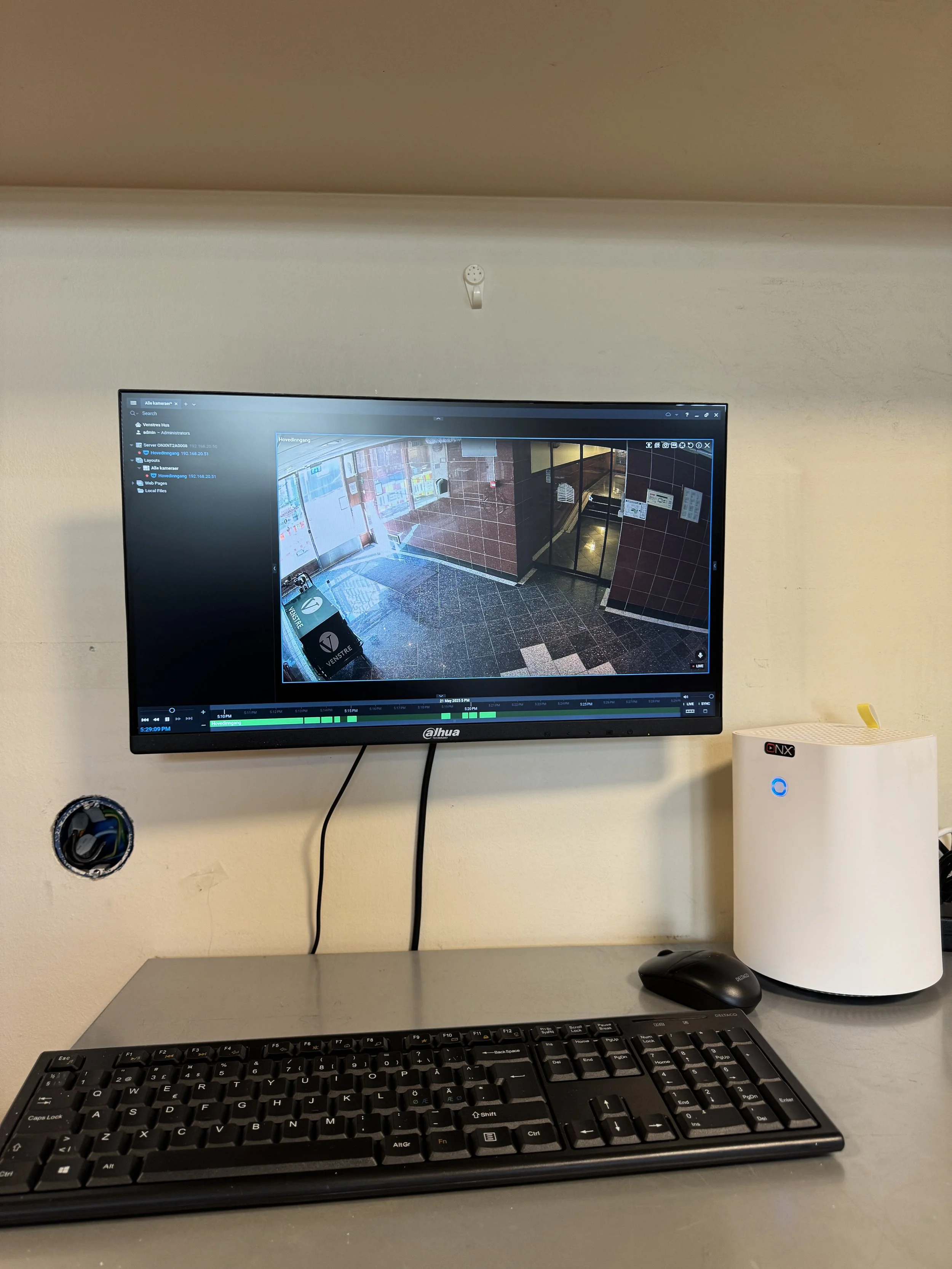 Kontor med en skrivebord, en svart PC-tastatur, en mus, en hvit sikkerhetsenhet og en veggmontert skjerm som viser et overvåkningskameraoverblikk.