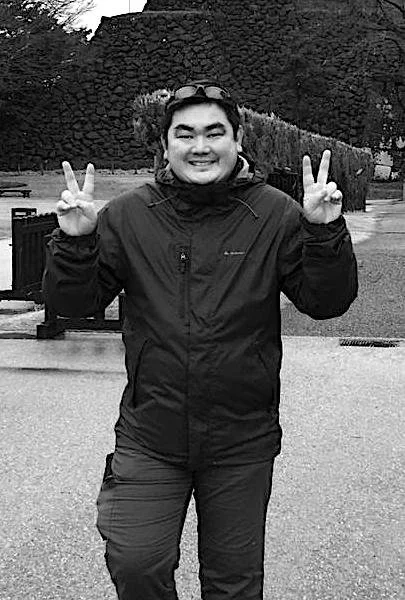 A person smiling and making peace signs with both hands outdoors in a park or garden setting, wearing a jacket and sunglasses on their head.