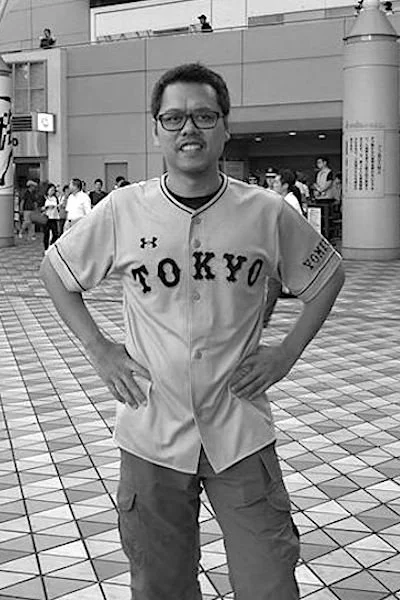 A man standing with hands on hips in a public space, wearing glasses and a baseball jersey with 'TOKYO' written on it, with other people in the background.