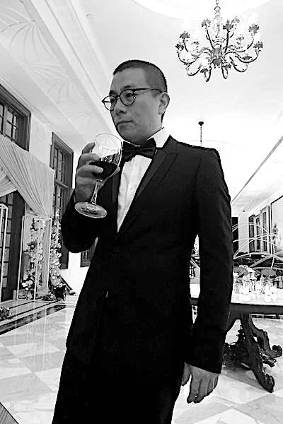 Man in tuxedo holding a glass of red wine in an elegant room with chandelier and large windows.