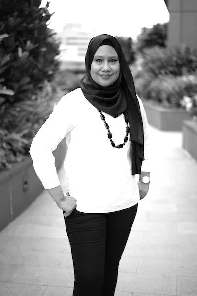 A woman wearing a hijab and white blouse standing outdoors on a city sidewalk.