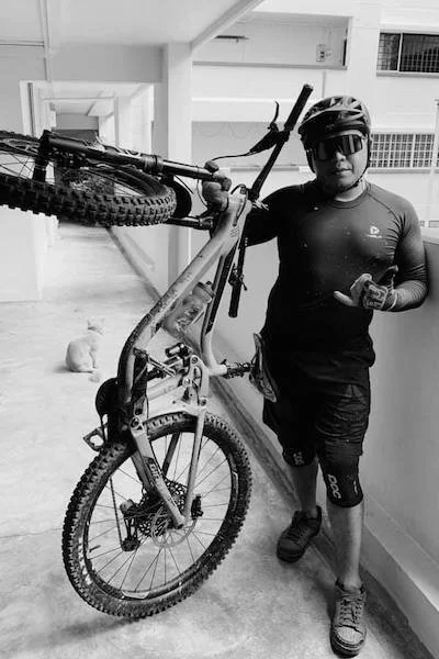 A man wearing a helmet, sunglasses, and cycling gear is standing next to a mountain bike, holding the handlebars and giving a thumbs-up, in an indoor corridor.